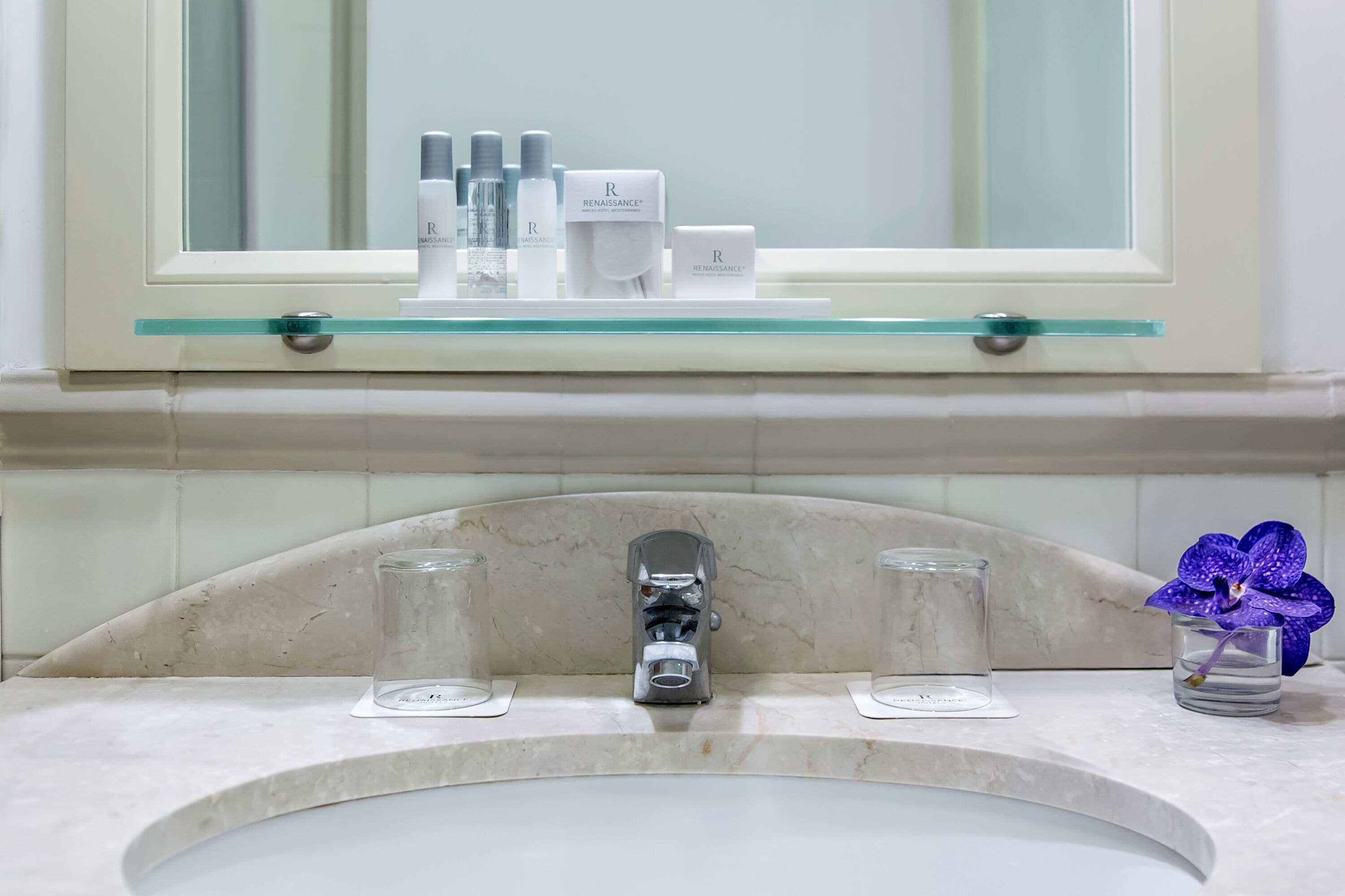 A bathroom with a marble sink, a glass shelf with toiletries, and a mirror above.