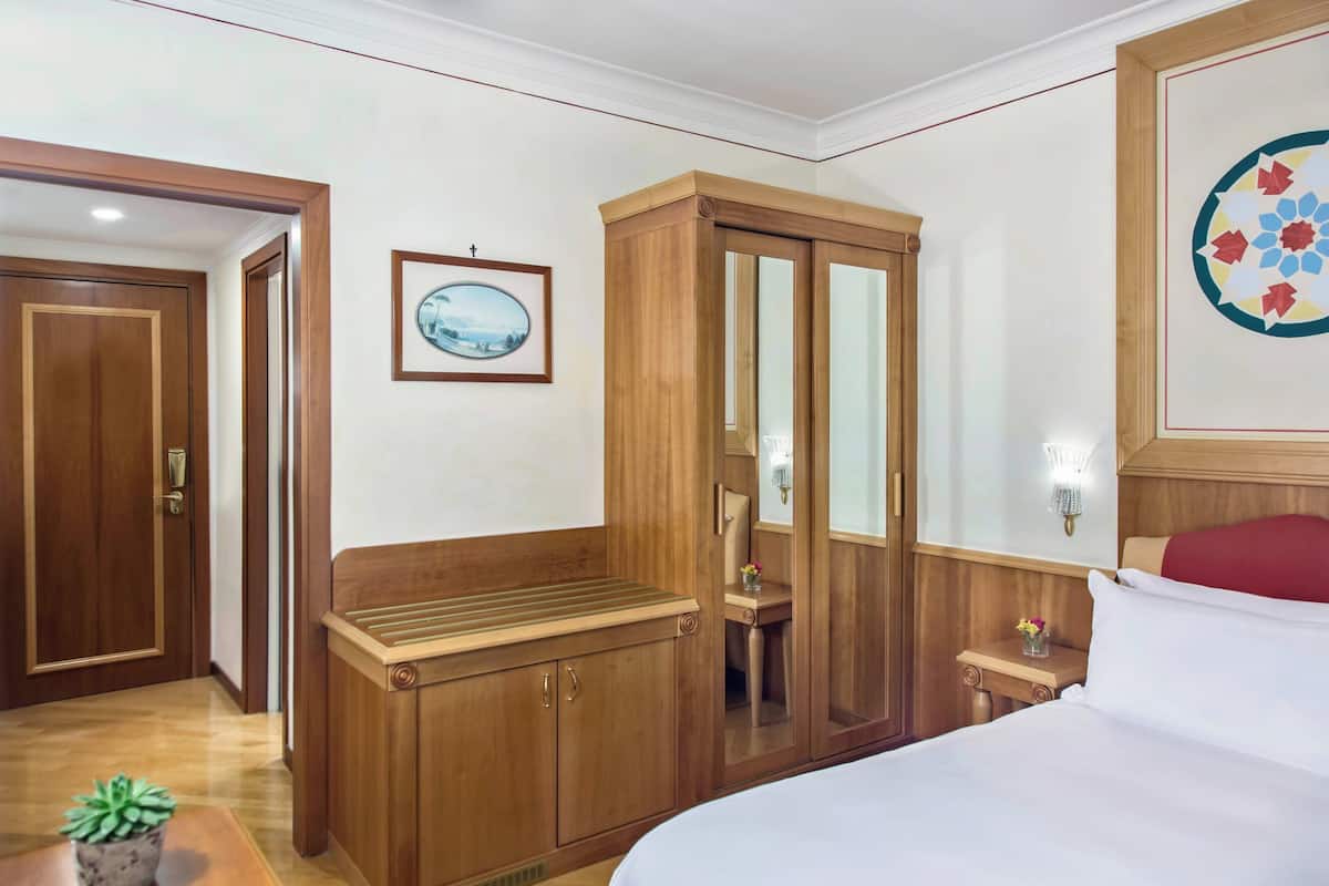 A hotel room with a wooden wardrobe, a bed with white bedding, a small table with a plant, and a framed picture on the wall.