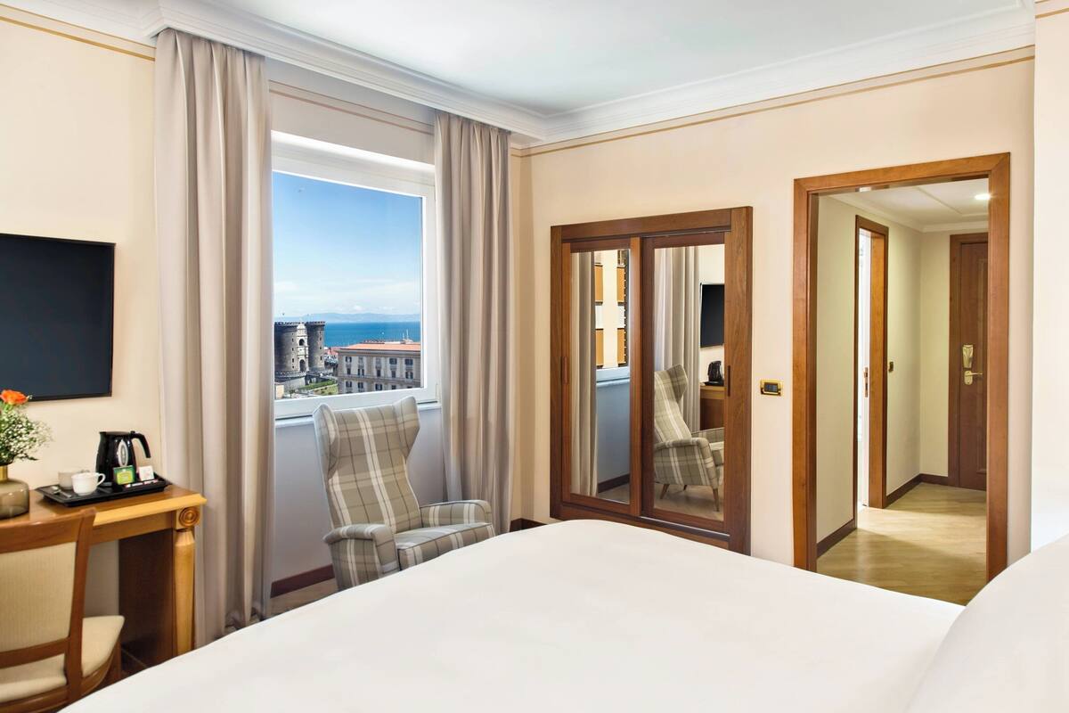 A hotel room with a bed, a desk with a coffee maker, a chair, and a large window with curtains.
