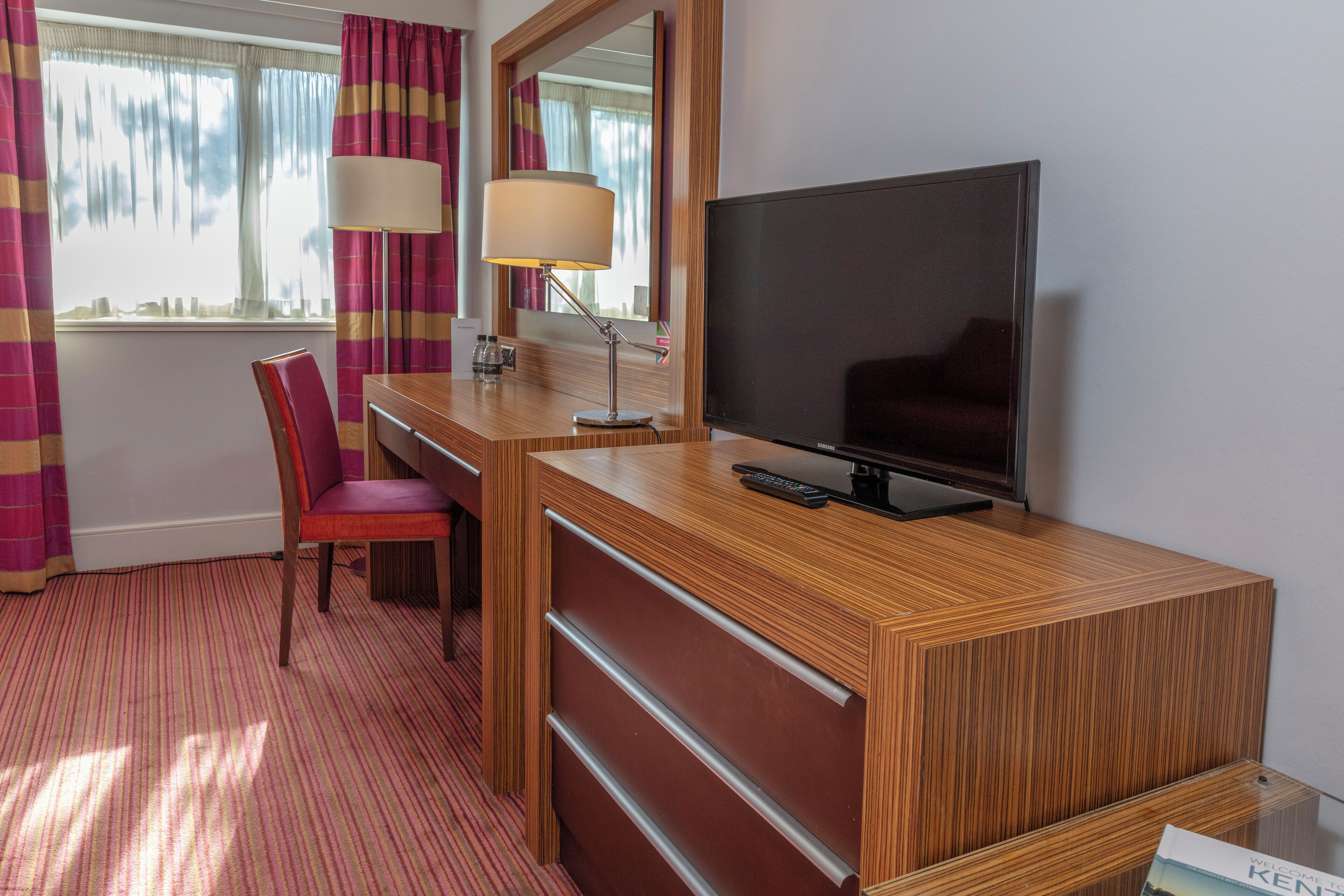 A hotel room with a wooden desk, a television, a chair, a lamp, and a window with curtains.