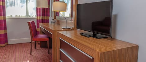A hotel room with a wooden desk, a television, a chair, a lamp, and a window with curtains.