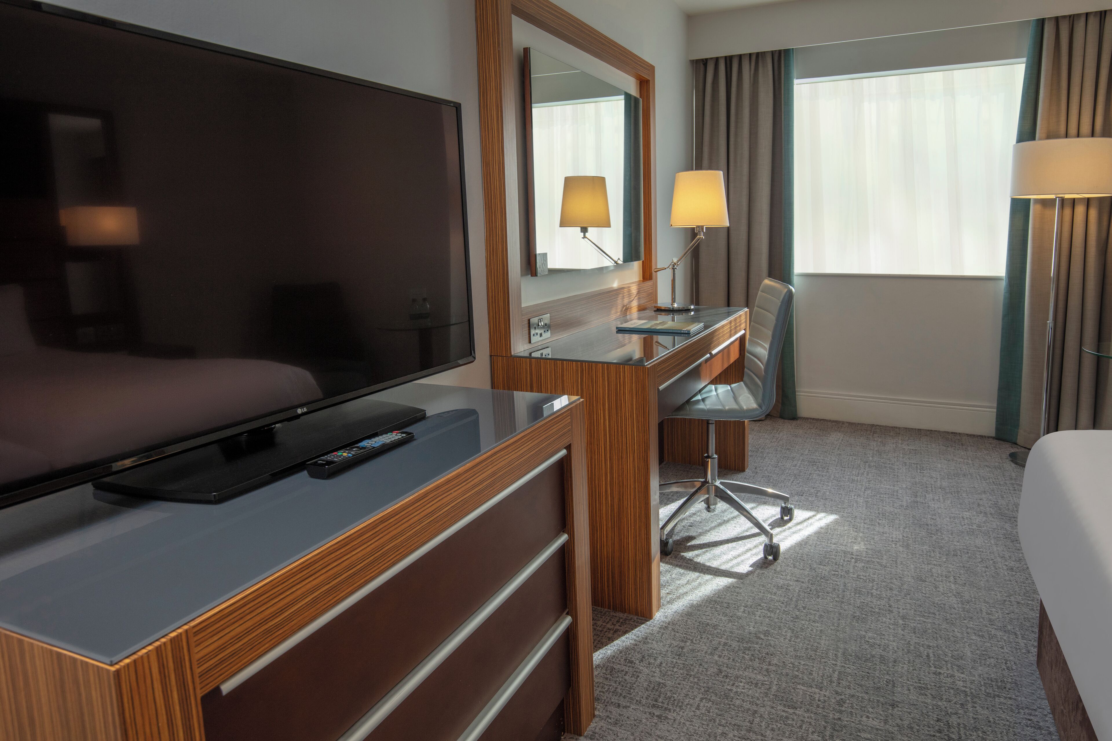A hotel room with a bed, a desk with a computer, a TV, a mirror, and a window with curtains.