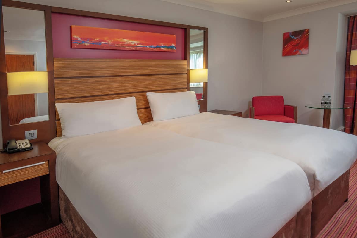 Une chambre d’hôtel avec un grand lit, une tête de lit en bois, une chaise rouge, une petite table avec une lampe et des œuvres d’art accrochées aux murs.