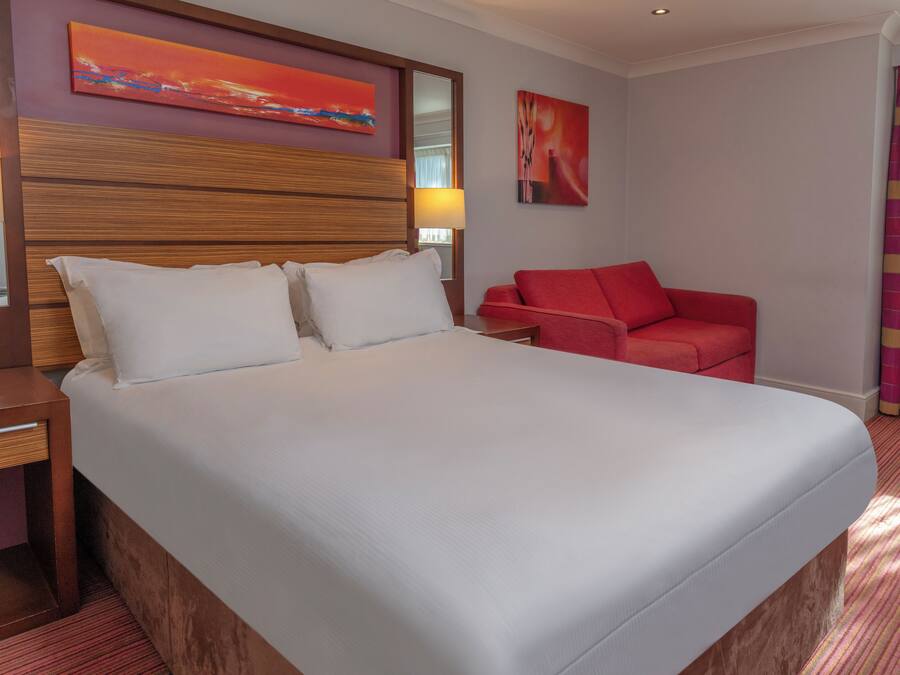 Ett hotellrum med en stor säng, en sänggavel i trä, en röd fåtölj och två tavlor på väggen.