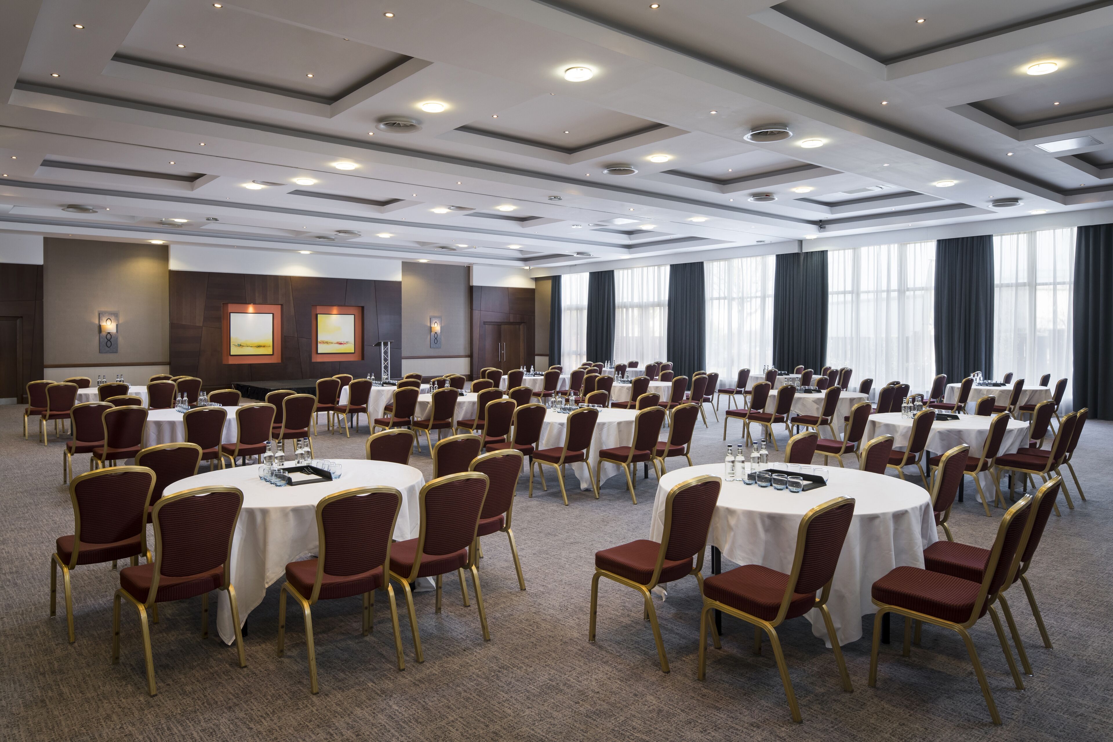 Une grande salle de conférence dotée de tables et de chaises rondes, équipée de luminaires encastrés et de grandes fenêtres.