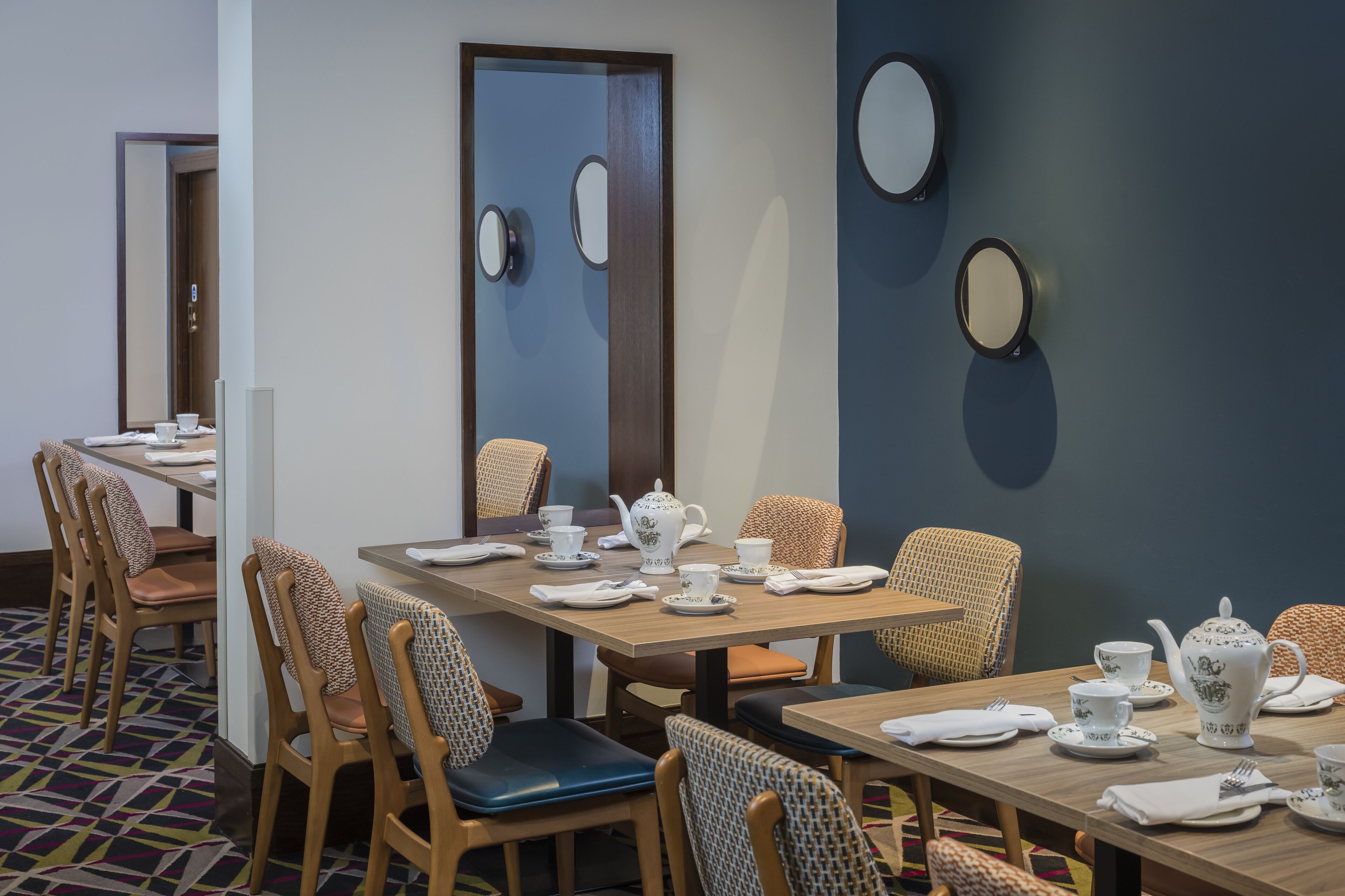 Een restaurantgedeelte in een hotel, met gedekte tafels voor een theeservies, stoelen met gestreepte kussens en decoratieve spiegelwanden.