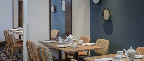 Un comedor de hotel con mesas dispuestas para el servicio de té, sillas con cojines estampados y espejos decorativos en la pared.