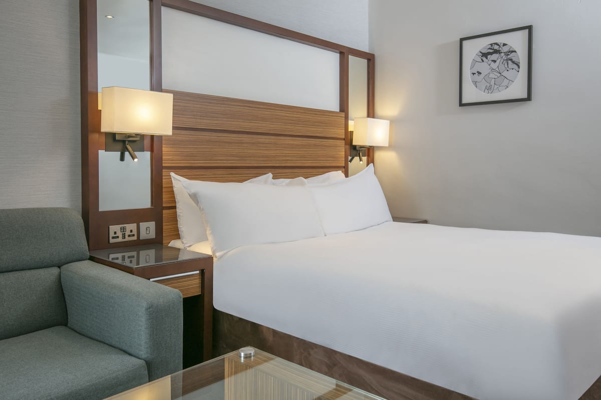 Une chambre d’hôtel avec un lit, une table de chevet, une lampe, une chaise et un tableau encadré au mur.