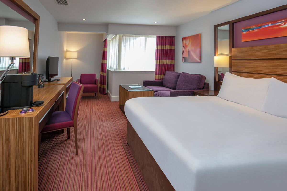 Une chambre d’hôtel avec un grand lit, un bureau avec un ordinateur, une chaise, un canapé, une petite table et une fenêtre avec des rideaux.