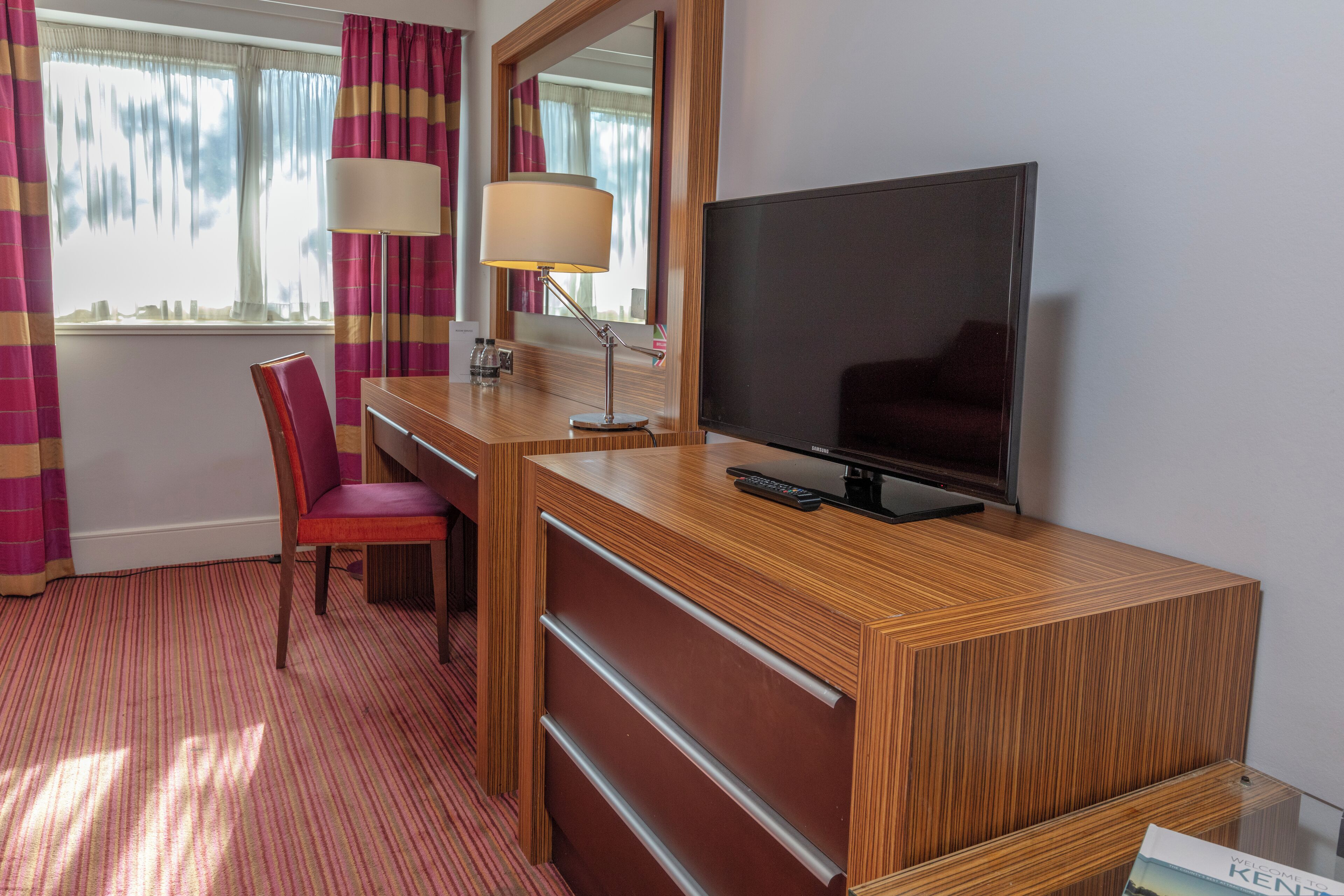 Quarto de hotel com escrivaninha de madeira, televisão, cadeira, abajur e janela com cortinas.