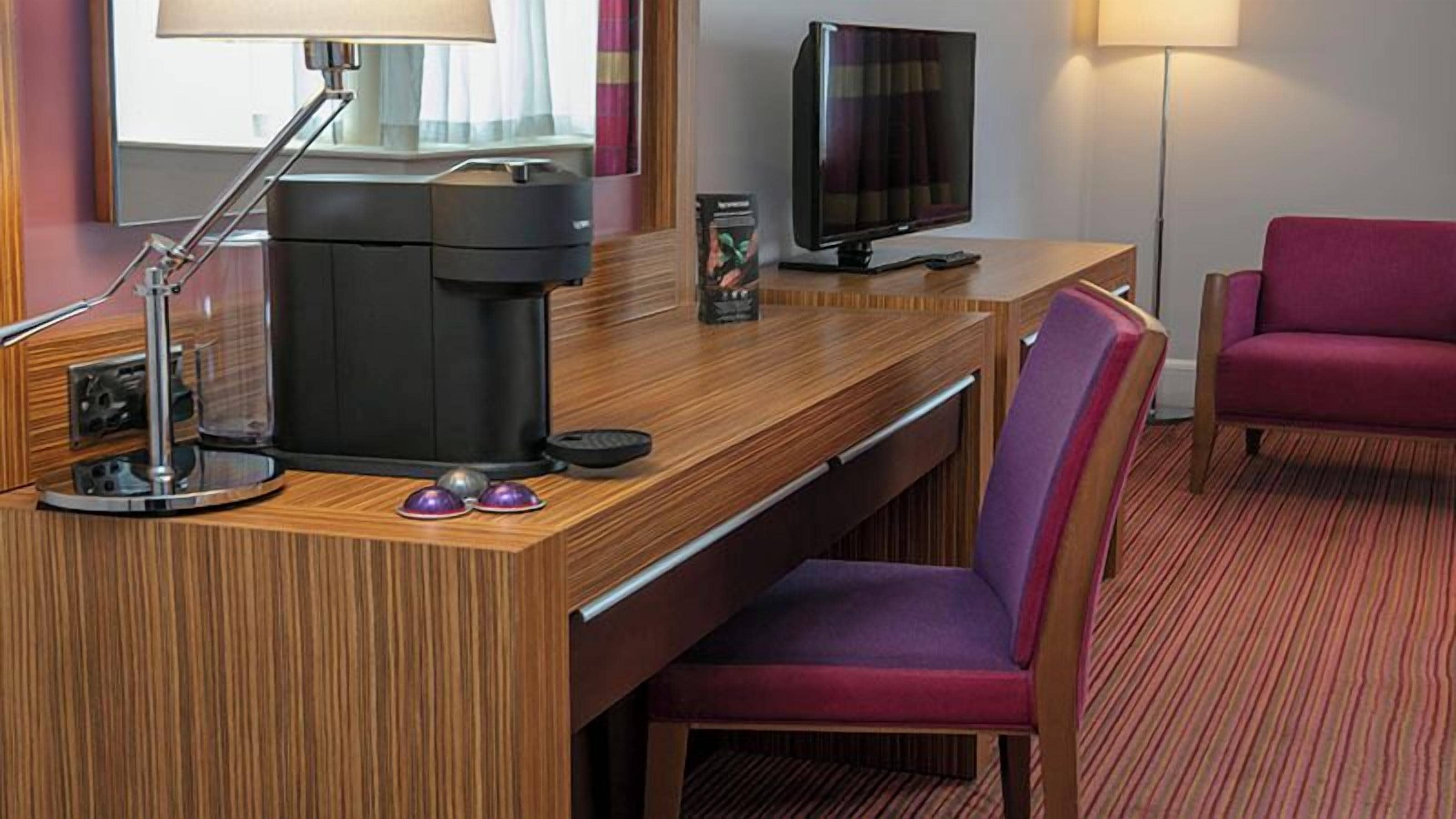 Hotelkamer met een houten bureau, een koffiezetapparaat, een televisie, een paarse stoel en een rood tapijt.
