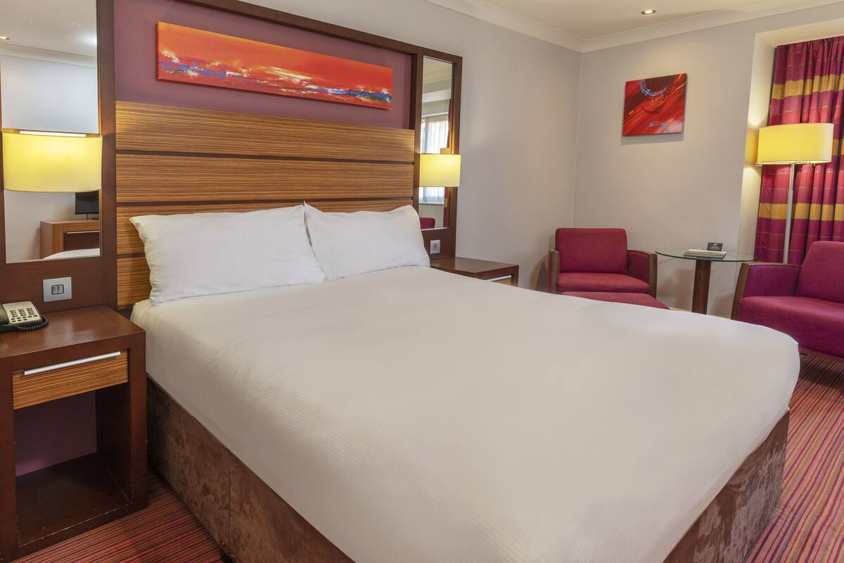 Une chambre d’hôtel avec un grand lit, une tête de lit en bois, une table de chevet avec un téléphone, une table en verre avec une chaise et deux lampes.