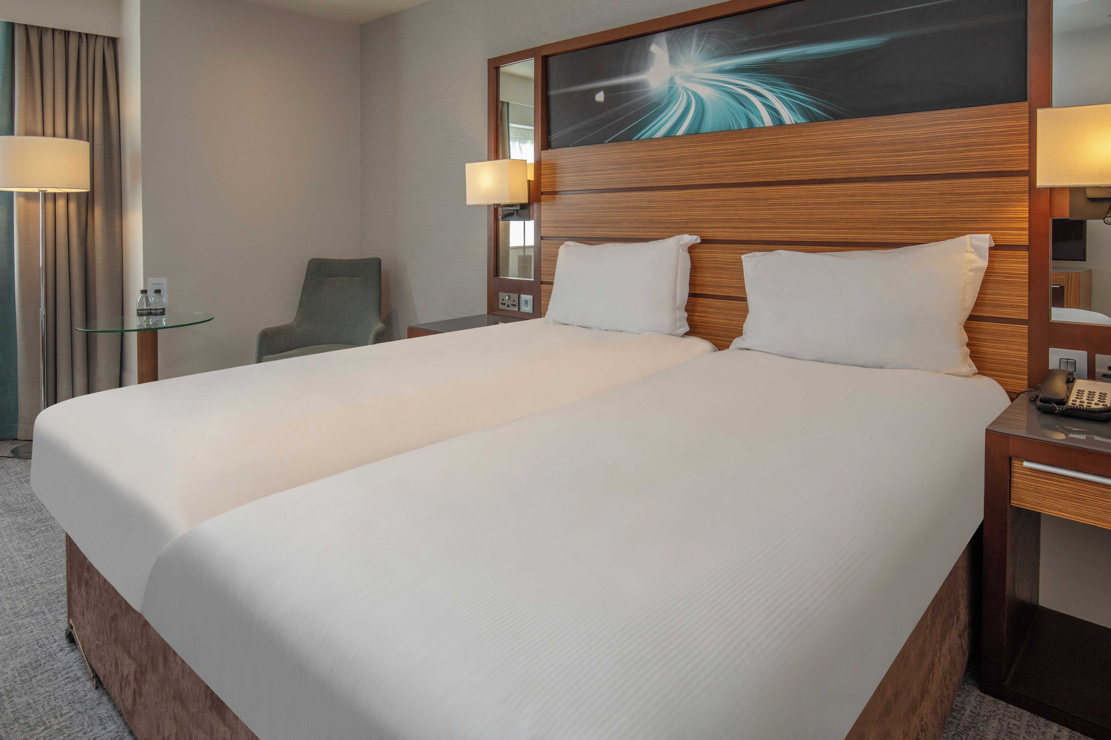A hotel room with a large bed, bedside lamps, a chair, and a glass table.