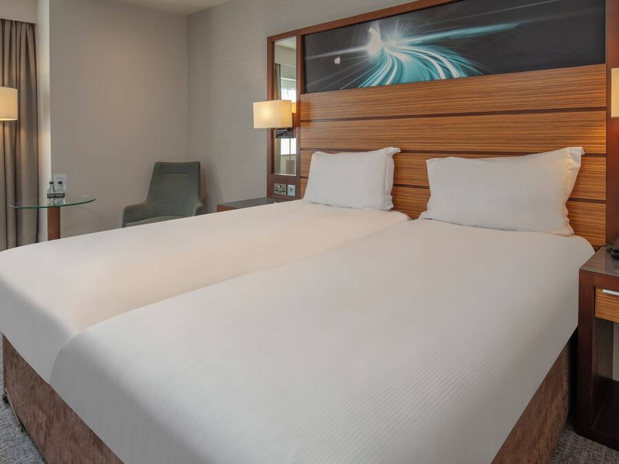 A hotel room with a large bed, bedside lamps, a chair, and a glass table.