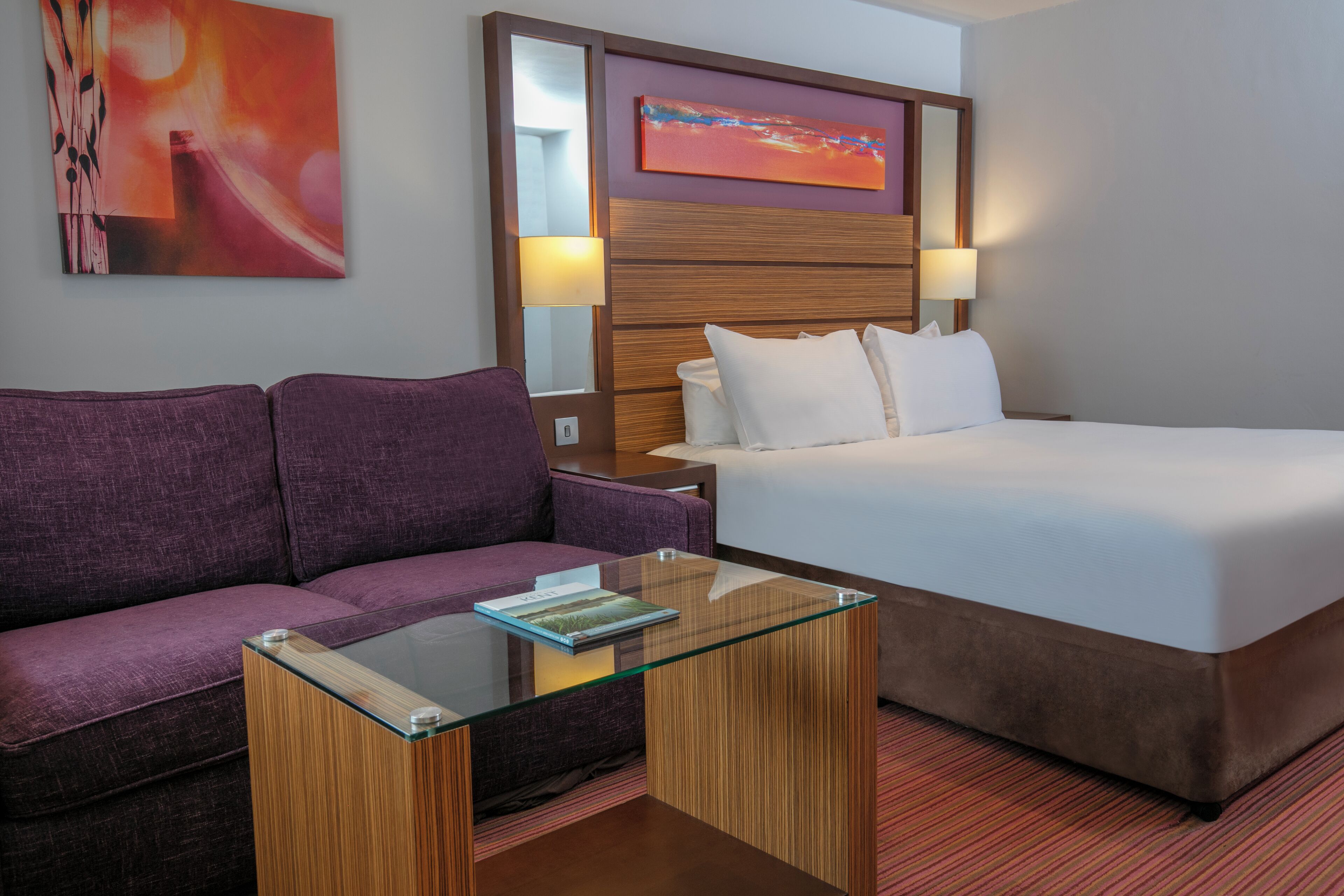 Ein Hotelzimmer mit einem Bett, einem violetten Sofa, einem Glastisch und zwei Wandleuchten.