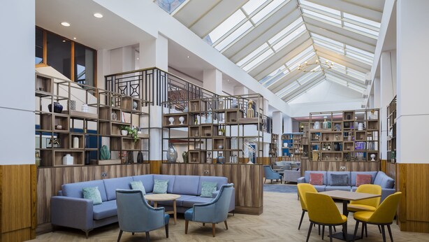 Een ruime leeshoek met een hoge plafond, houten boekenplanken, een blauwe bank en gele stoelen.