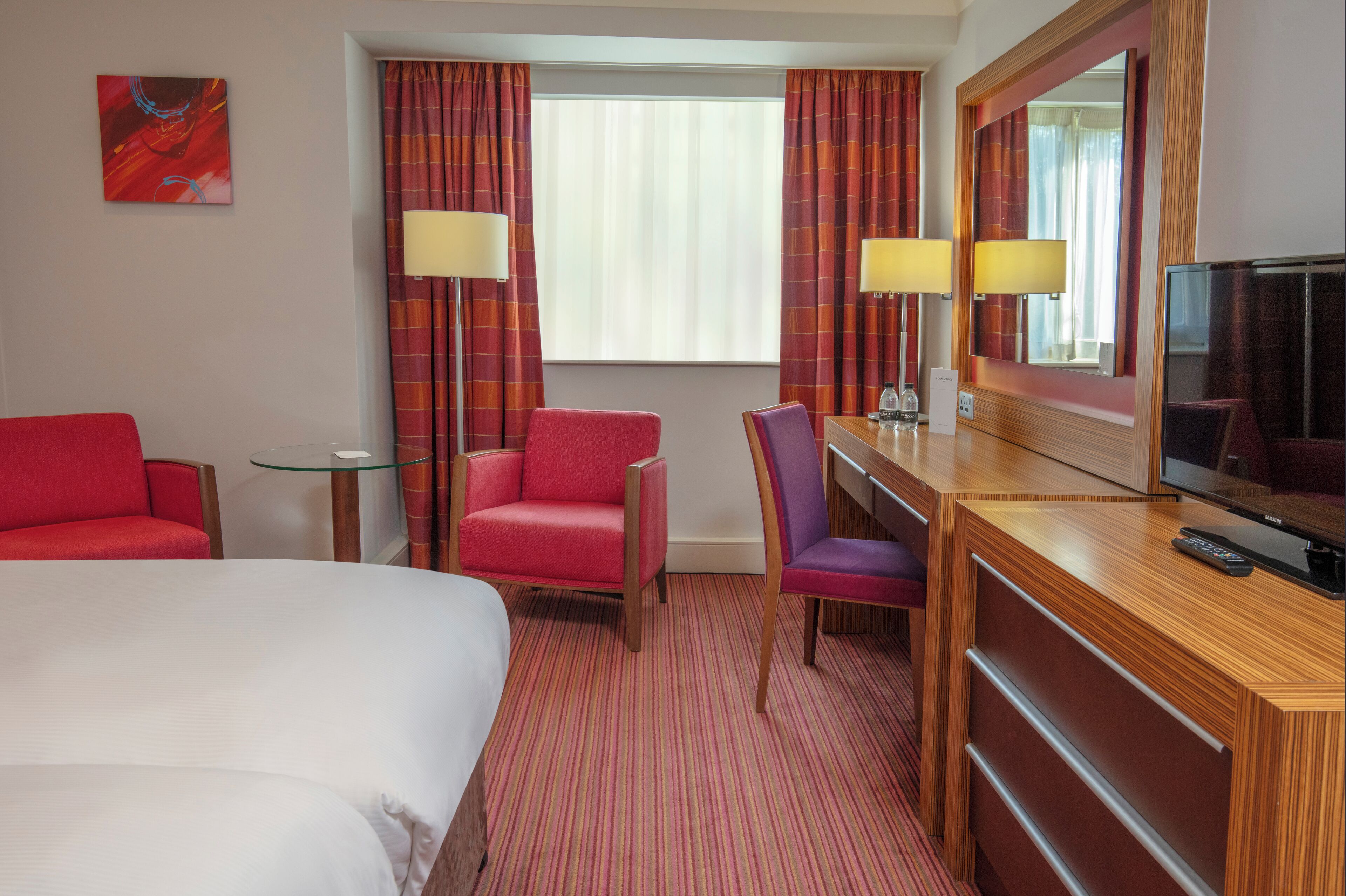 A hotel room with a bed, a desk with a TV, two chairs, a small table, and a window with curtains.