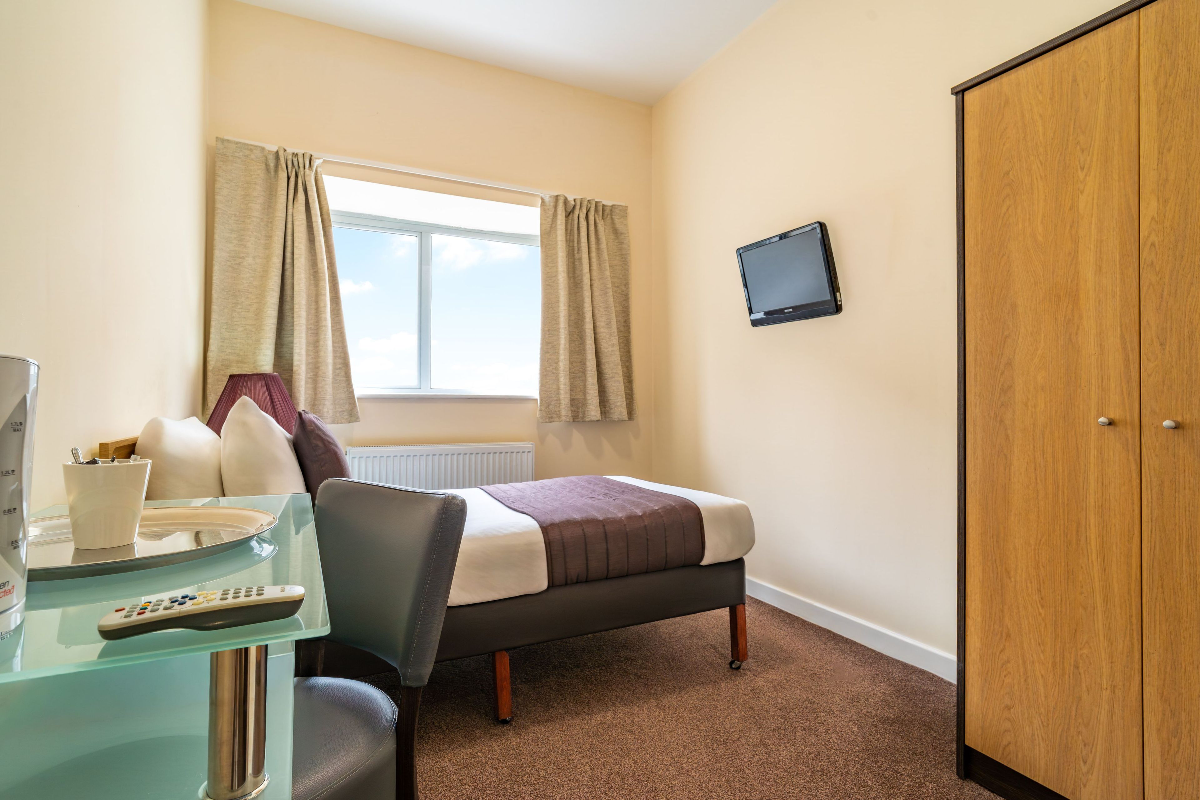 A hotel room with a bed, a desk with a lamp, a television on the wall, and a wardrobe.