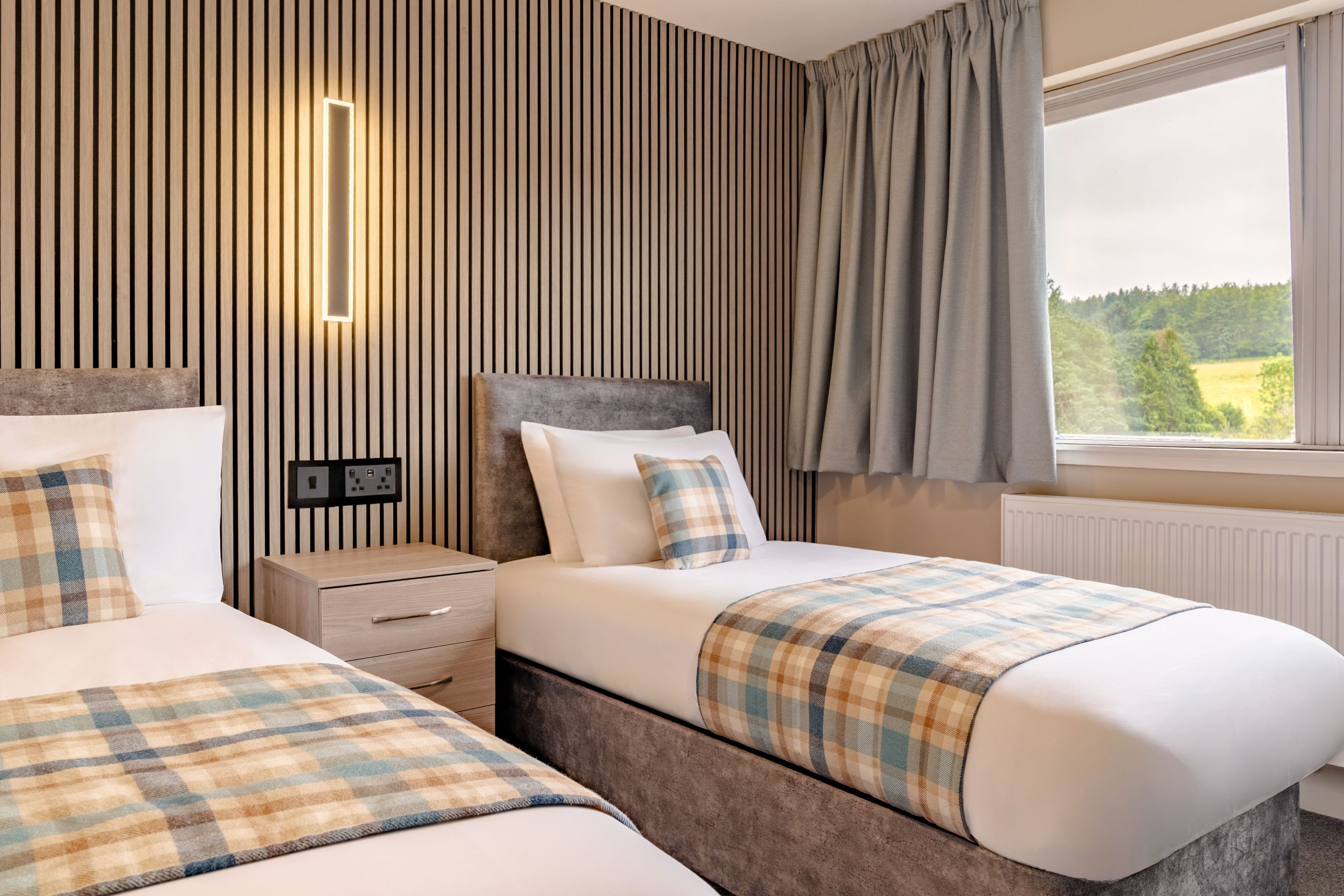 A hotel room with two beds, a nightstand, a dresser, a window with curtains, and a view of the outdoors.