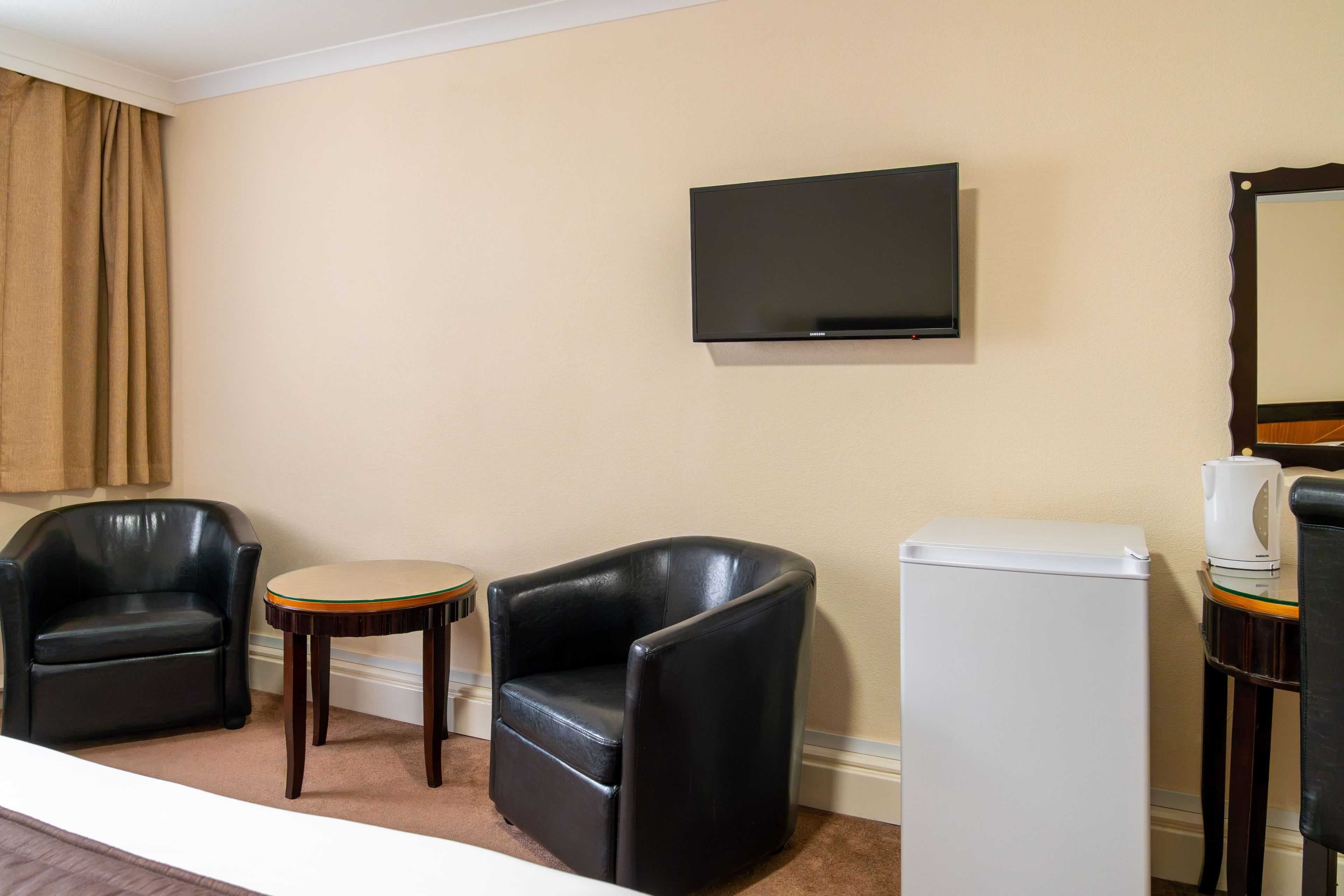Une chambre d’hôtel avec deux fauteuils en cuir noir, une petite table ronde, un téléviseur à écran plat fixé au mur, un réfrigérateur blanc et une machine à café posée sur une table d’appoint.