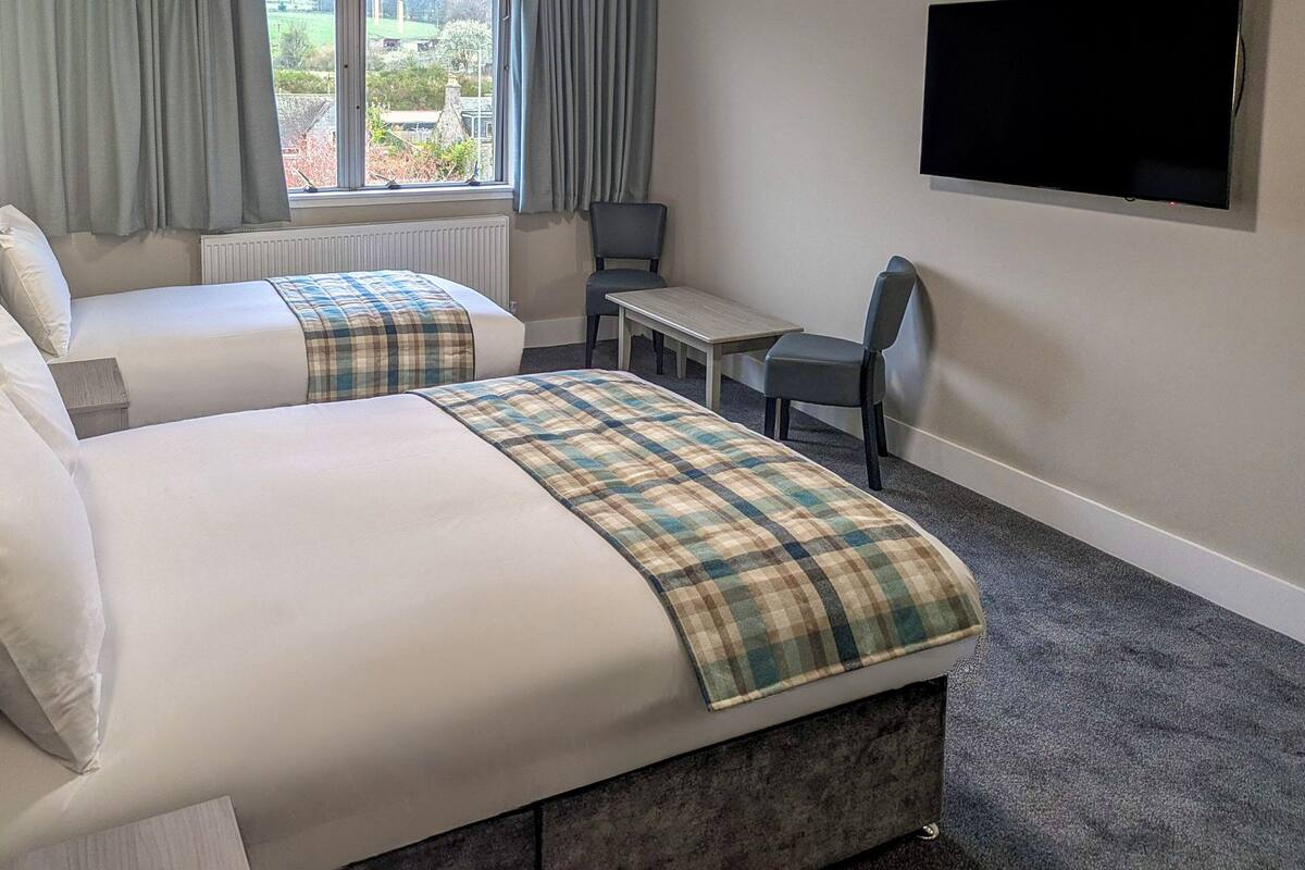 Habitación de hotel con dos camas, una mesita, una silla, un televisor y una ventana con cortinas.