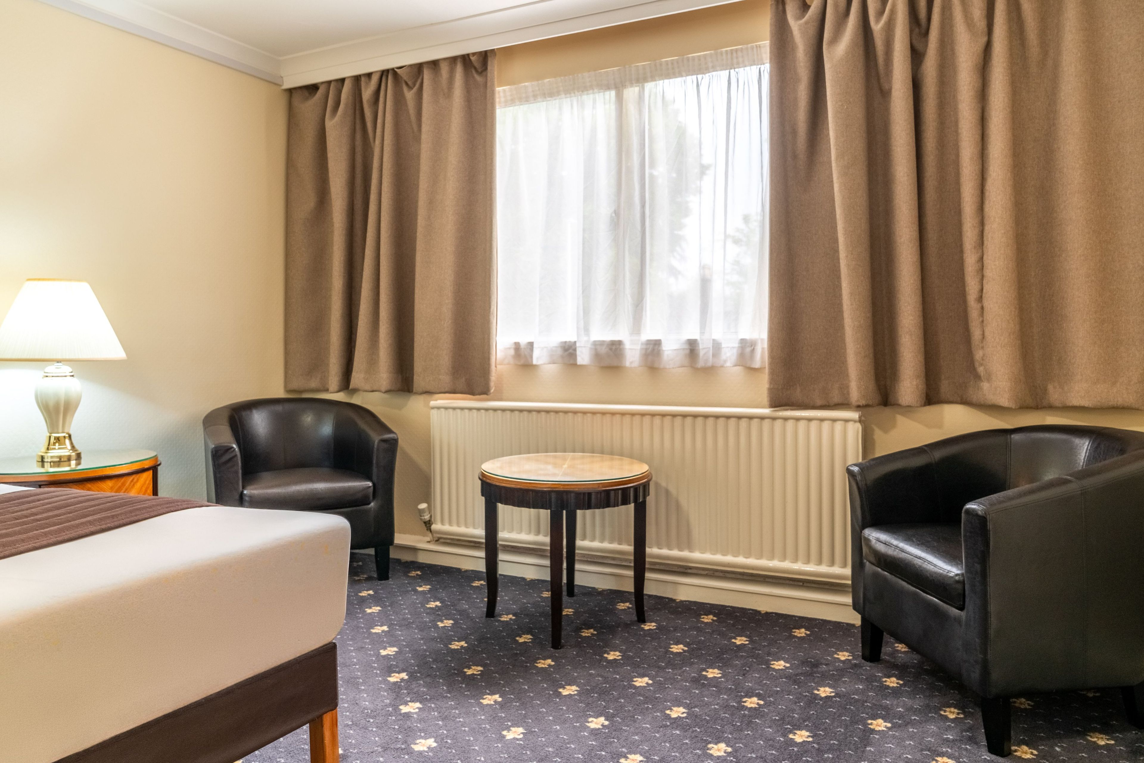 A hotel room with a bed, two armchairs, a small table, a lamp, and a window with curtains.