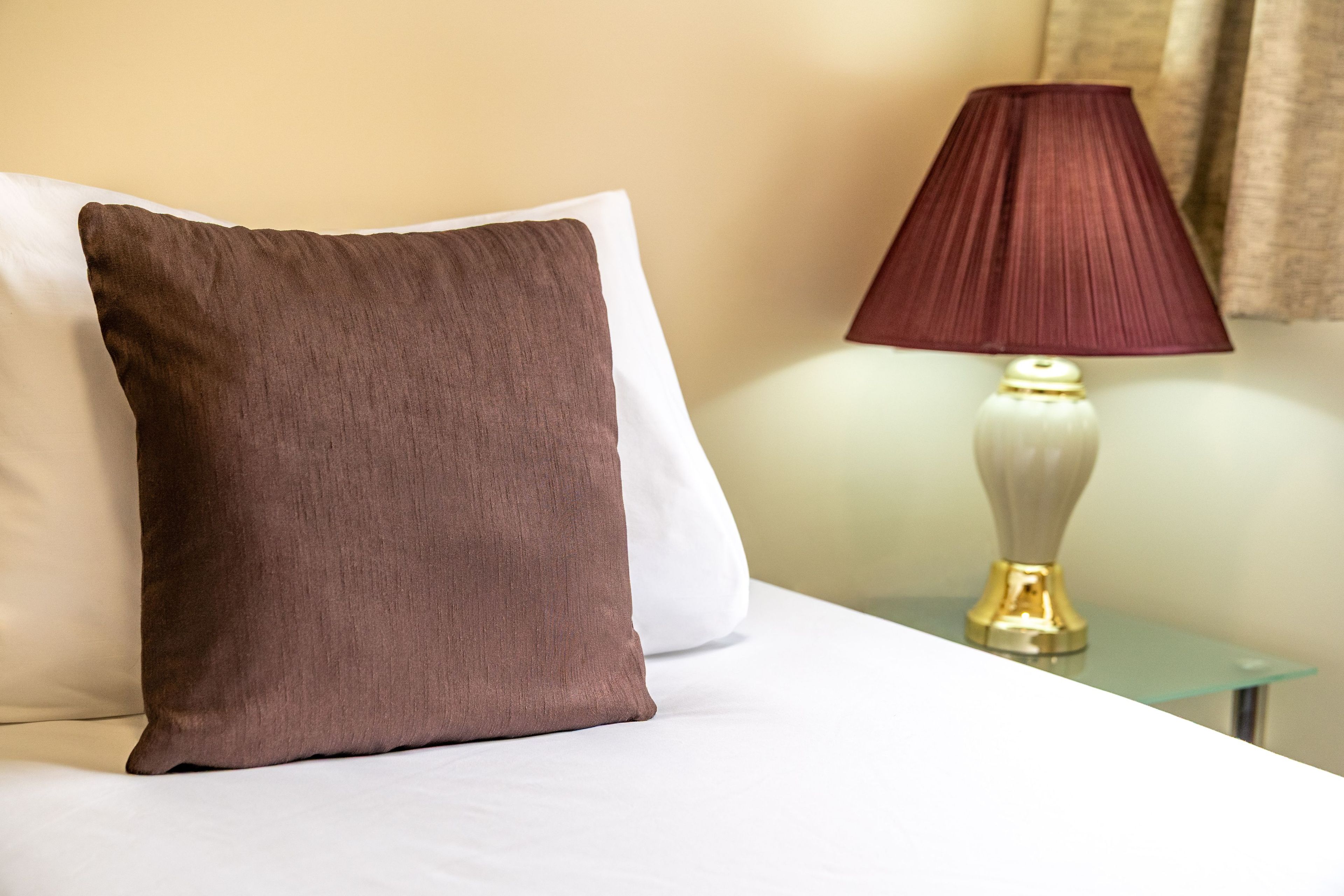 Ein Bett mit einem weißen Kopfkissen und einem braunen Kissen, eine Nachttischlampe mit rotem Schirm und ein kleiner Tisch mit einer Lampe.