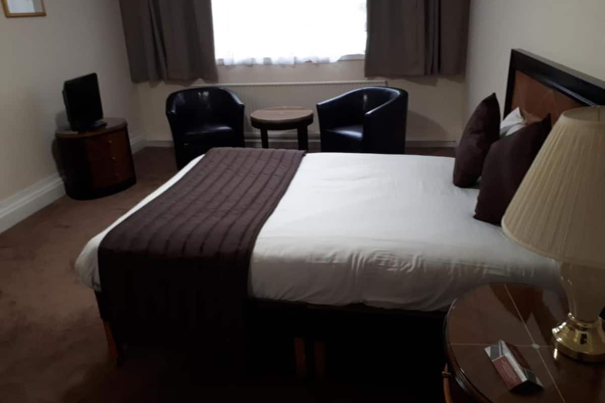 Habitación de hotel con una cama grande, una mesita de noche con lámpara, una pequeña mesa con sillas y un altavoz sobre un mueble.