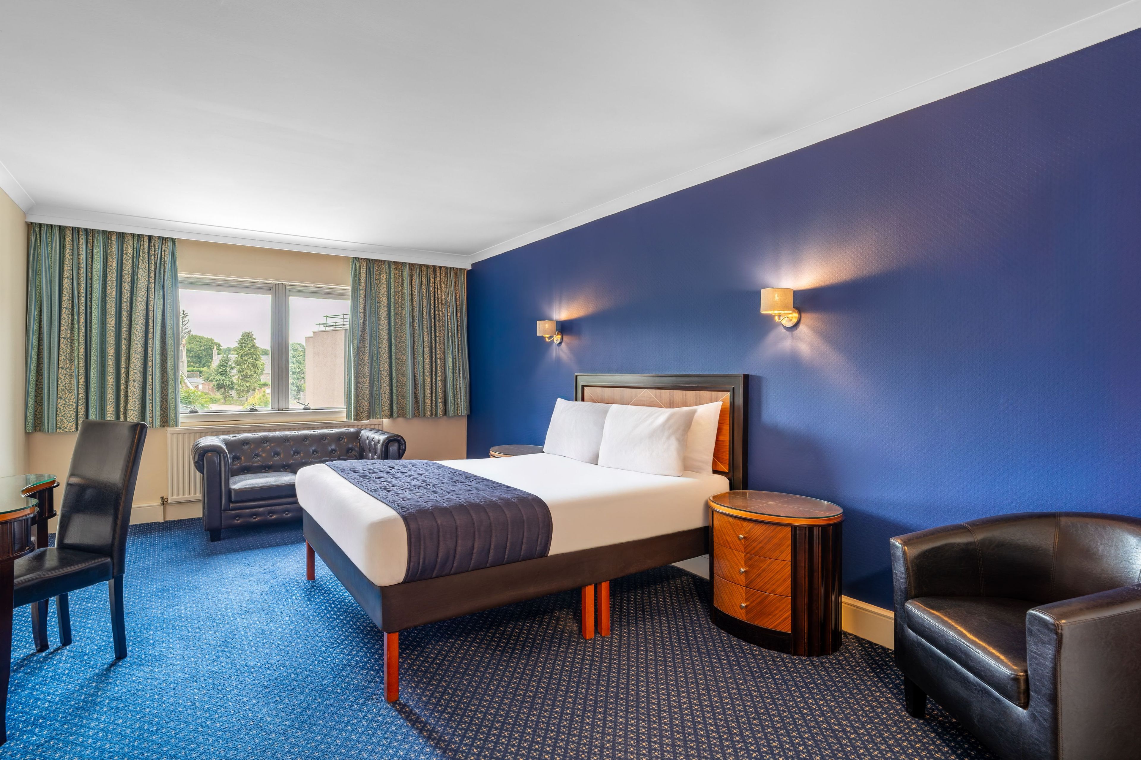 Ein Hotelzimmer mit einem großen Bett, einer Sitzecke mit Stuhl und Tisch sowie Blick ins Freie durch das Fenster.