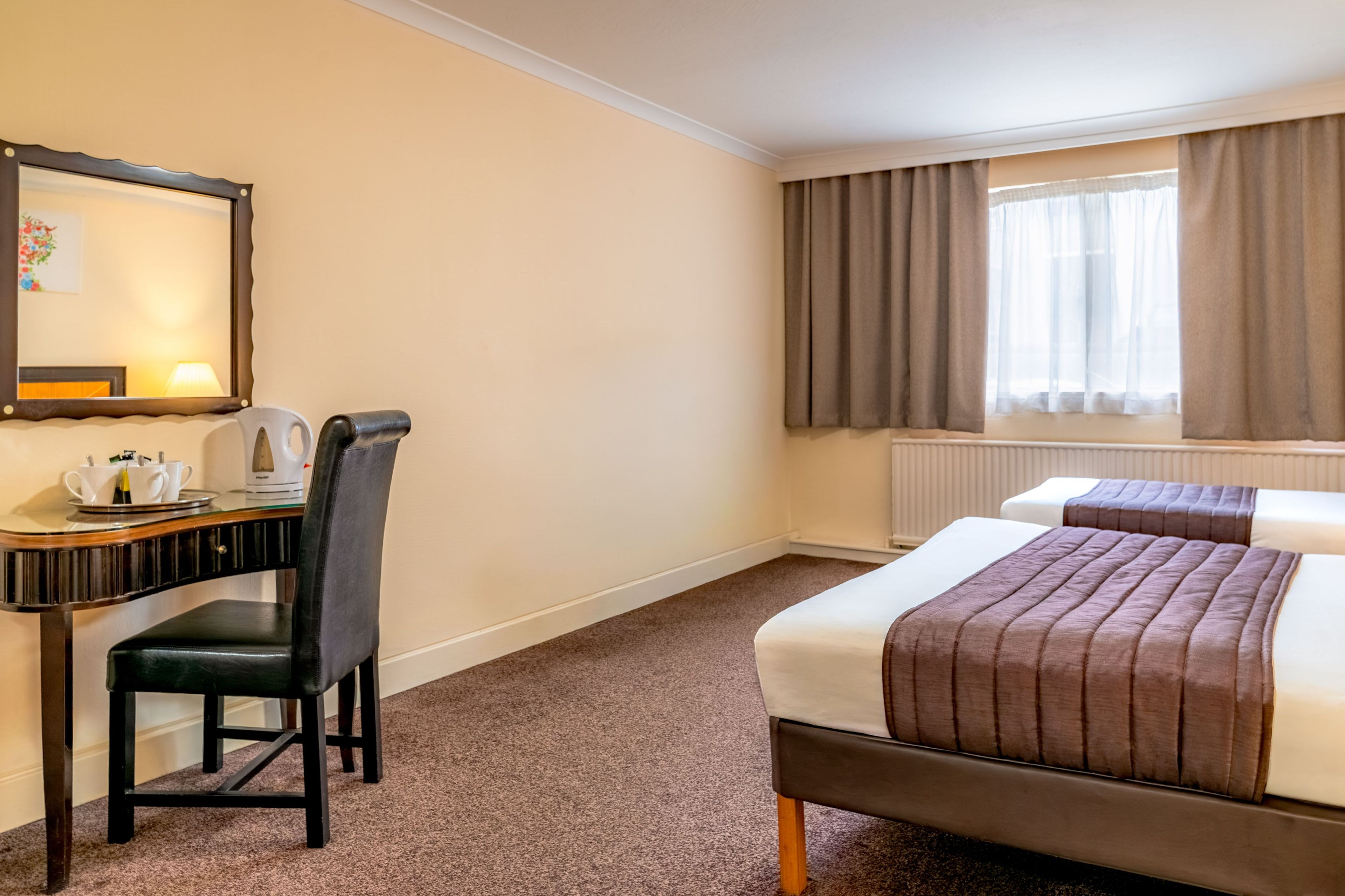 A hotel room with a desk, chair, two beds, a window with curtains, and a radiator.