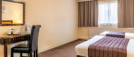 A hotel room with a desk, chair, two beds, a window with curtains, and a radiator.