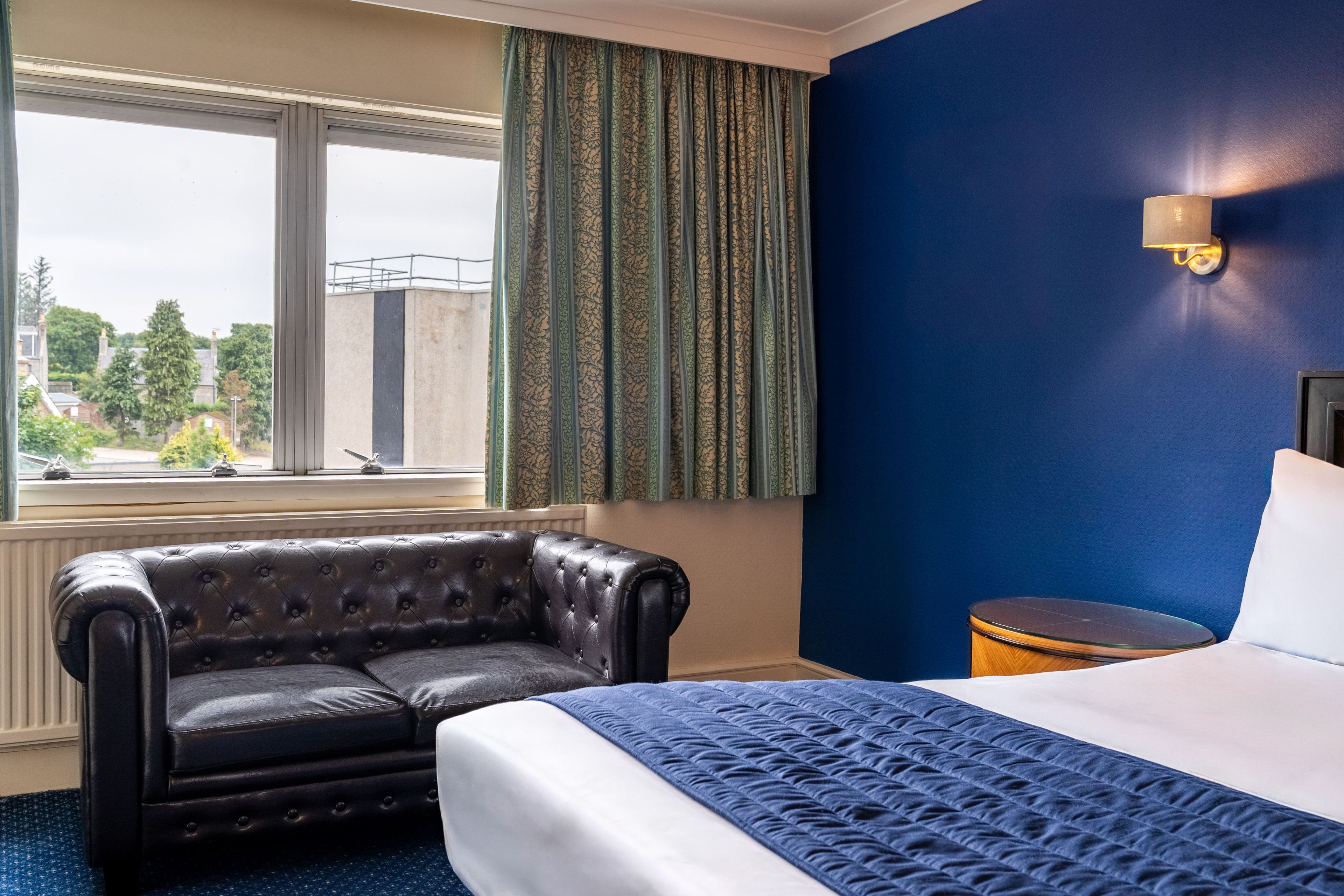 A hotel room with a bed, a leather armchair, a nightstand, and a window with curtains.