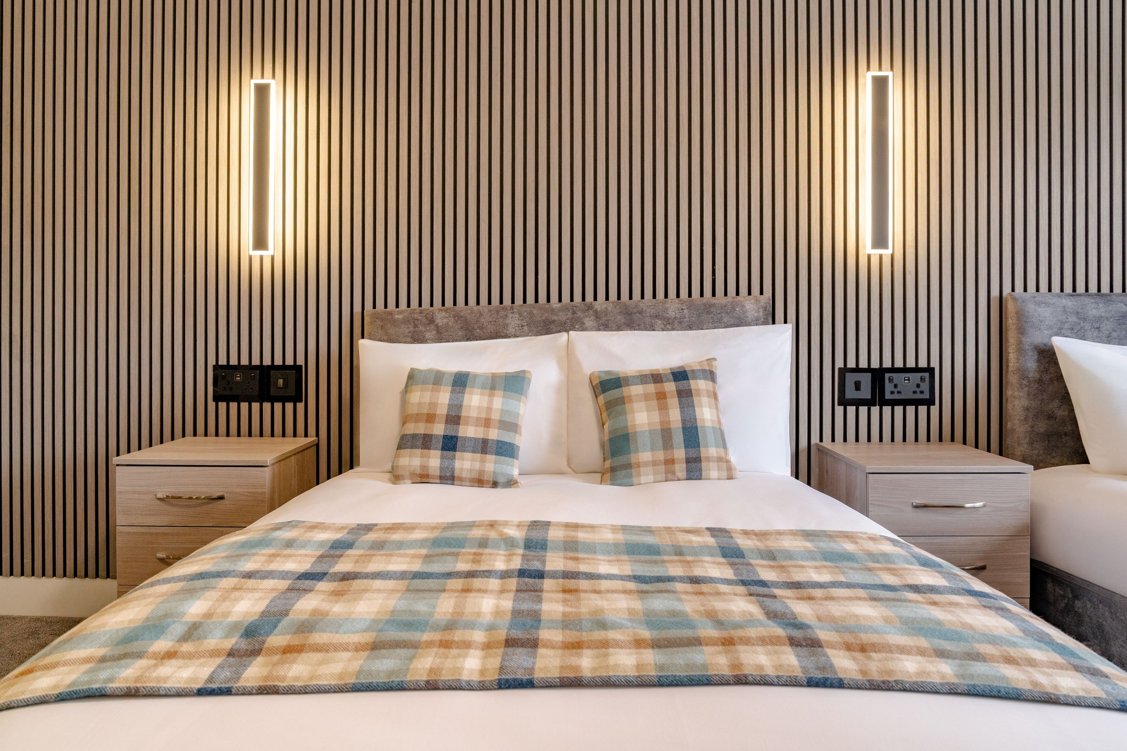 A hotel room with a large bed, two bedside tables, a headboard, and wall-mounted lighting.