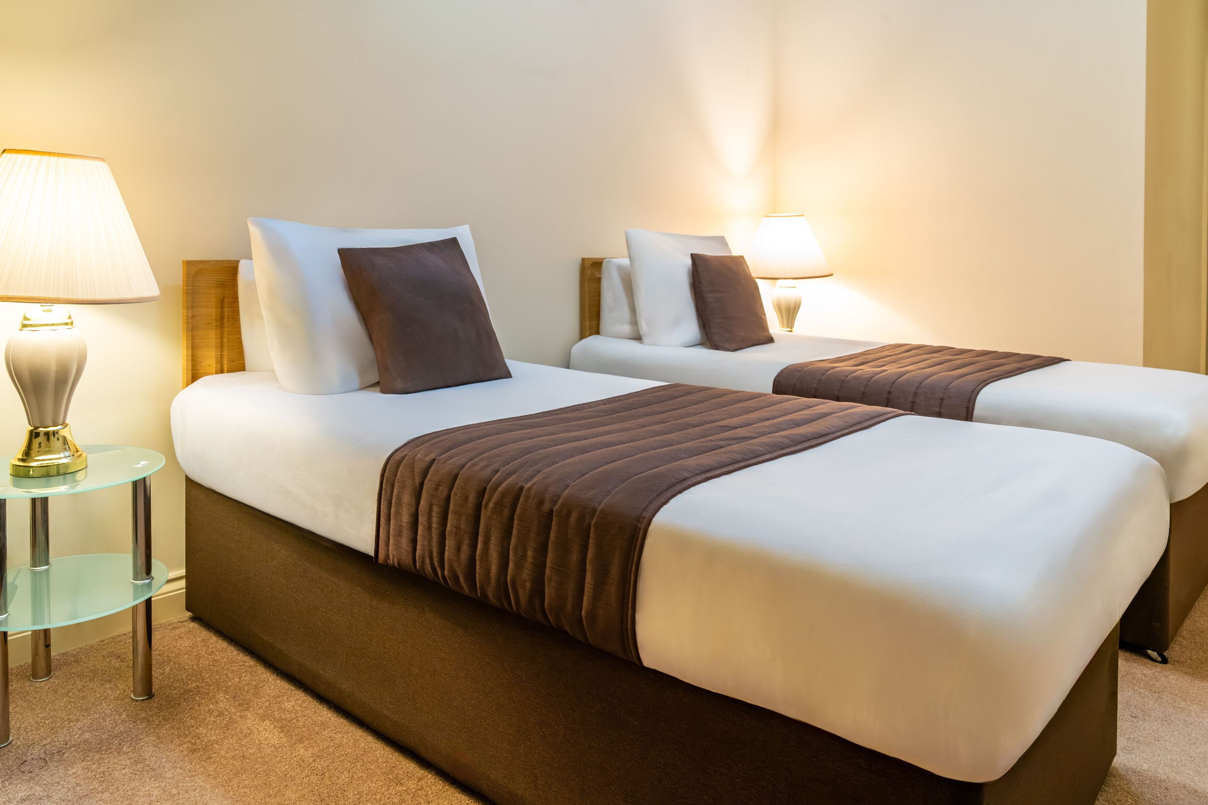 Ein Hotelzimmer mit zwei Betten, beide mit weißen Leinentüchern und braunen Kopfteilen. An jeder Seite der Betten befinden sich Nachttischlampen.