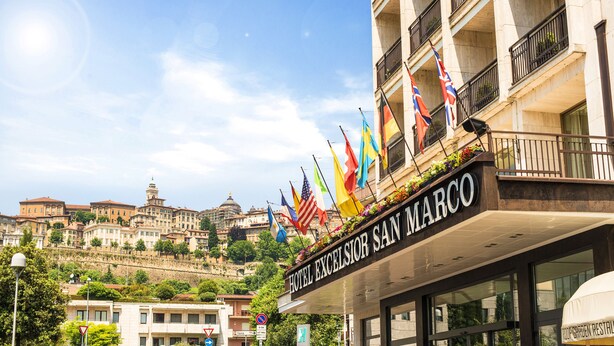 The Hotel Excelsior San Marco with multiple flags, a view of historic buildings in the background, and a clear sky. - Hotel Excelsior San Marco (Bergamo)