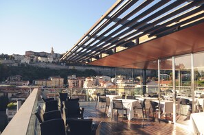 A rooftop terrace with tables and chairs, offering a view of a historic cityscape.