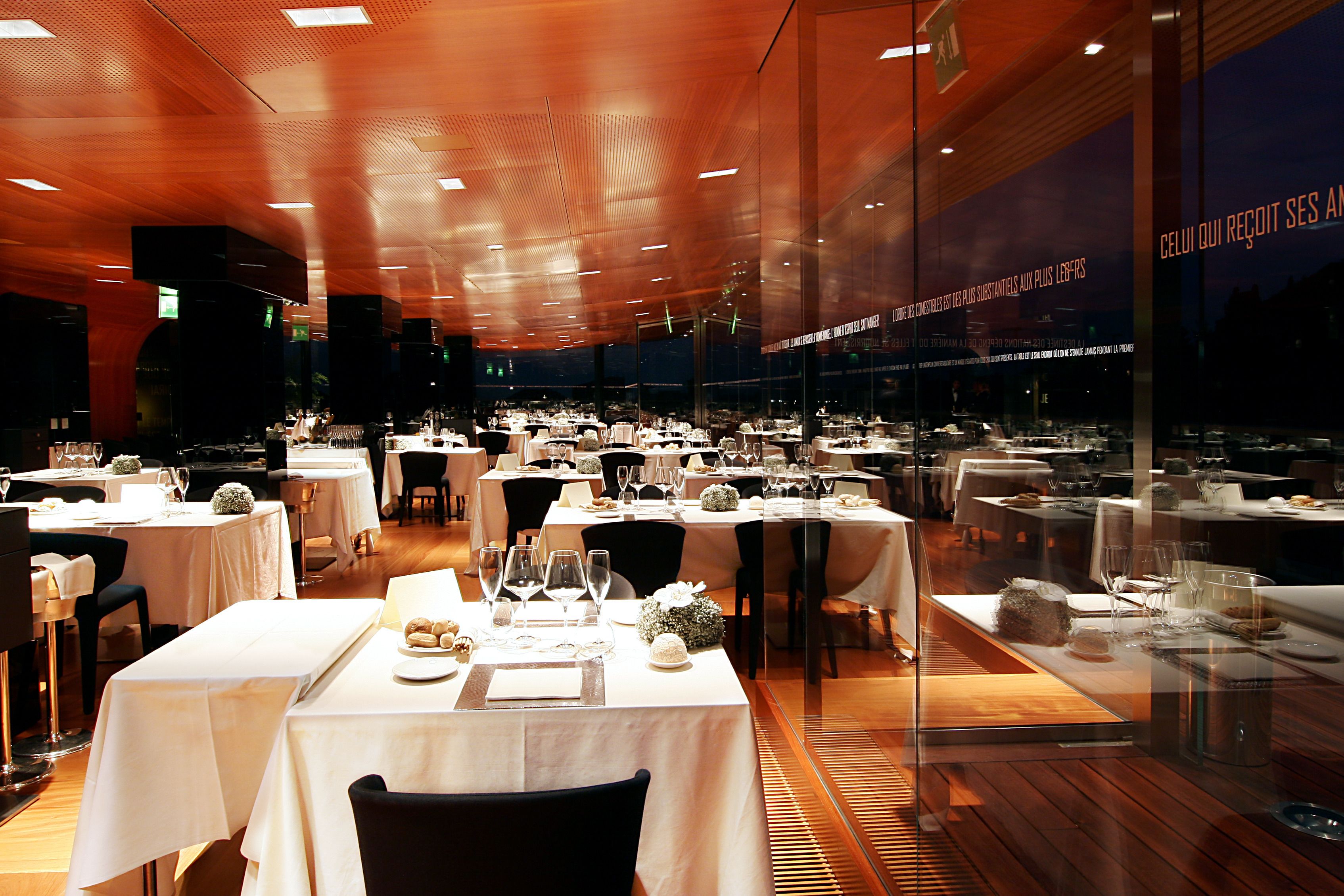 Un restaurante con varias mesas preparadas para comer, con una decoración moderna que incluye paneles de techo de madera y divisiones de vidrio.