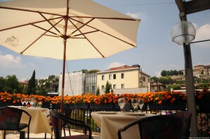 An outdoor dining area with tables and chairs, a large umbrella, and a view of buildings and trees in the background.