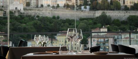 A dining table with empty wine glasses and plates, set against a backdrop of a cityscape with historic buildings and a stone wall.