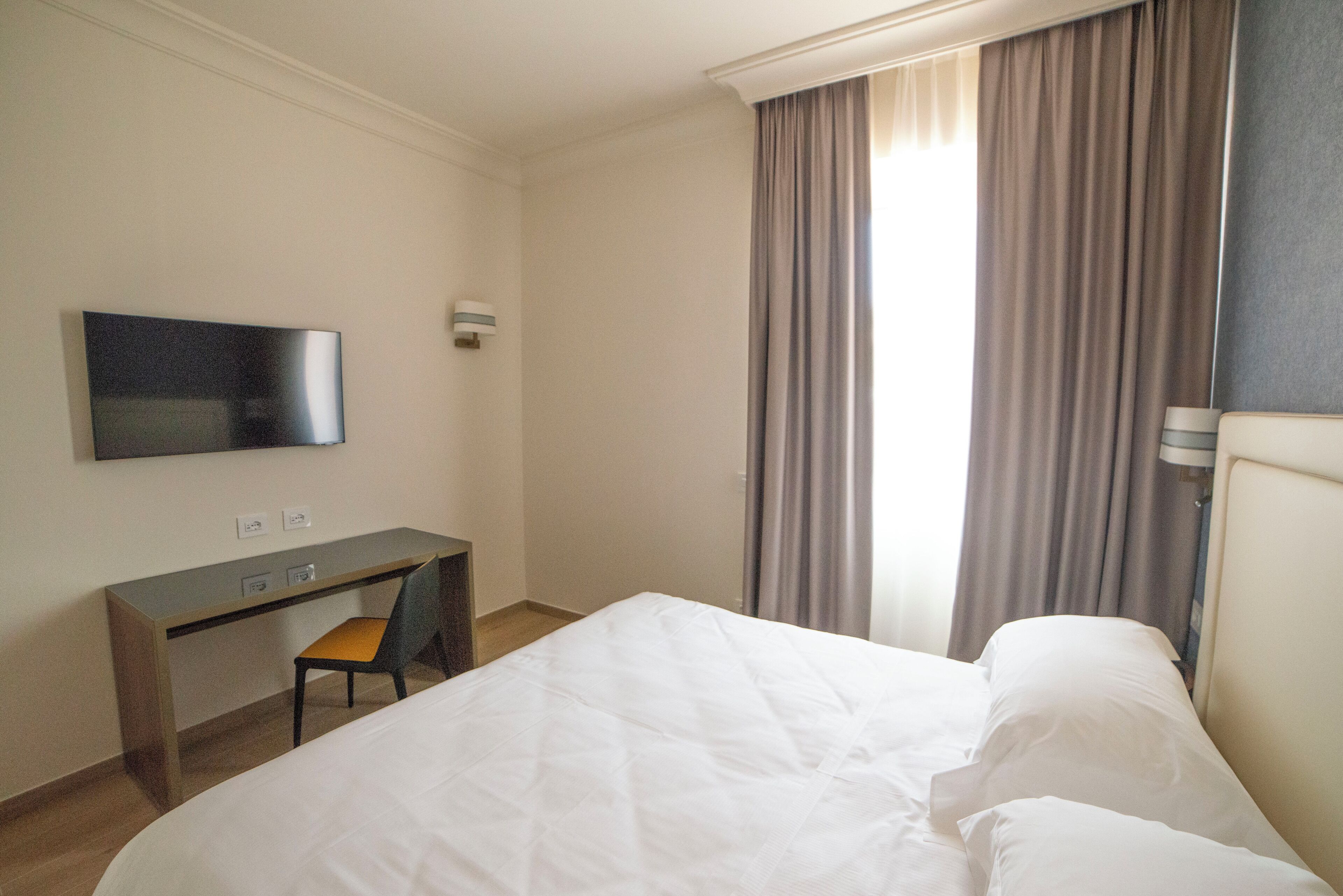 A hotel room with a bed, a desk, a chair, a TV, and a window with curtains.