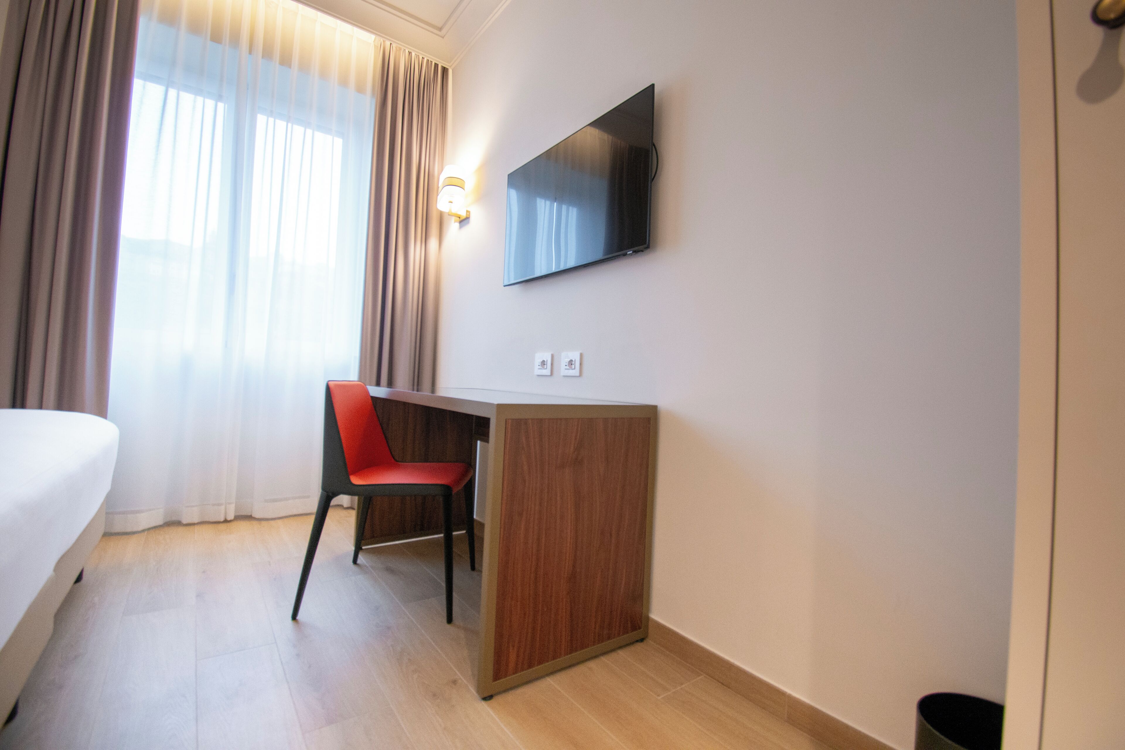 Habitación de hotel con cama, escritorio con silla, televisor montado en la pared y ventana con cortinas.
