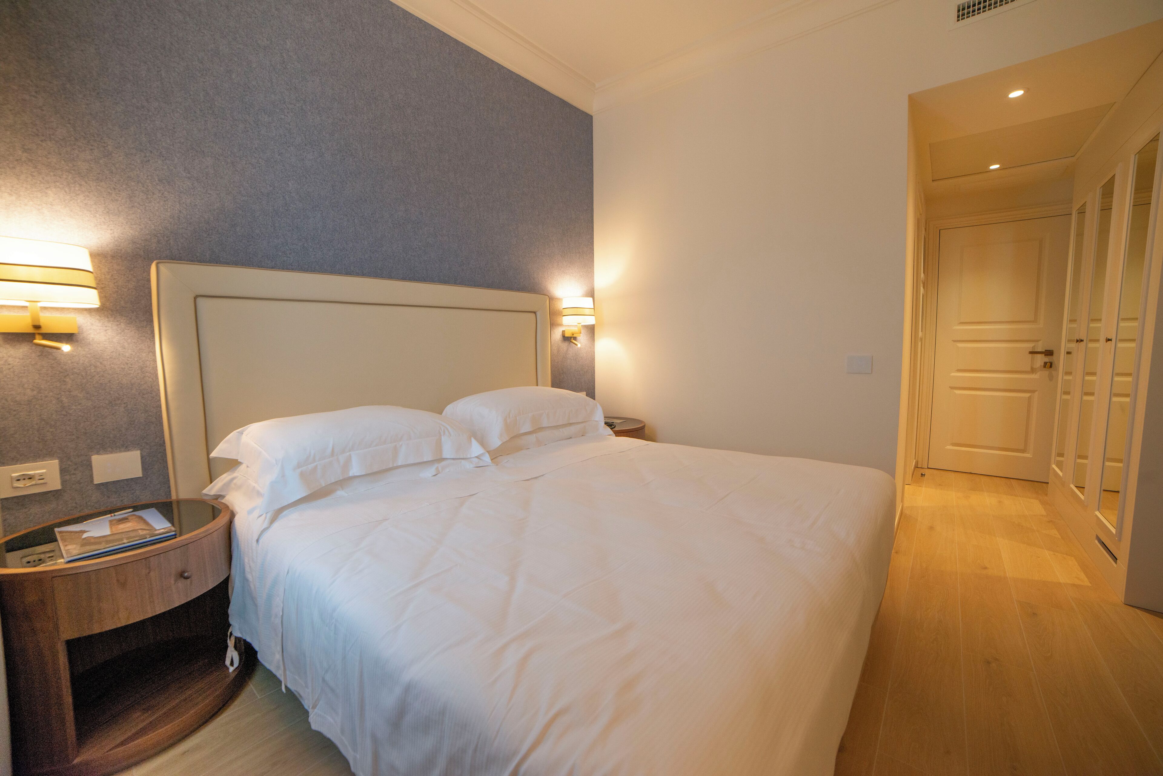 A hotel room with a large bed, bedside lamps, a wooden nightstand, and a door leading to another room.