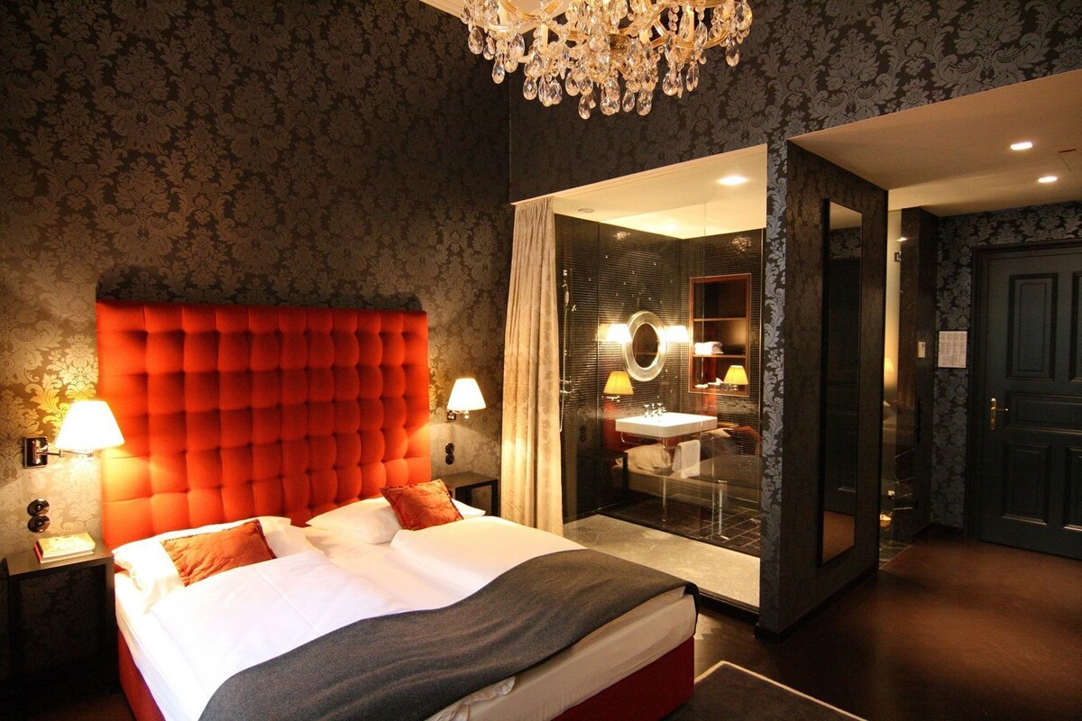 A hotel room with a tufted headboard, a large bed, a glass-enclosed bathroom, and a chandelier.