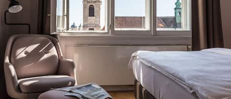A hotel room with a bed, a chair, a table with a newspaper, a lamp, and a window with a view of a clock tower.