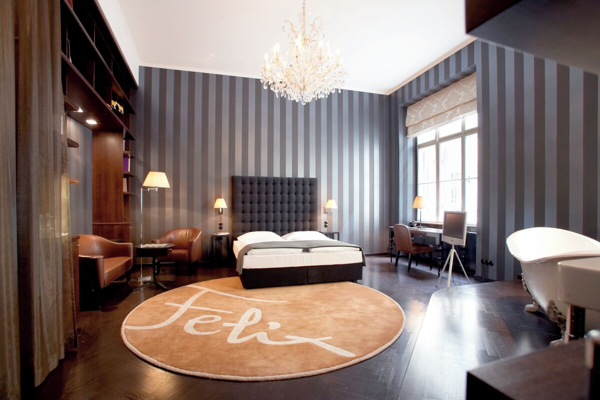 A modern hotel room with a tufted headboard, a round rug with the word 'Felic,' a chandelier, a desk with a chair, and a bathtub.