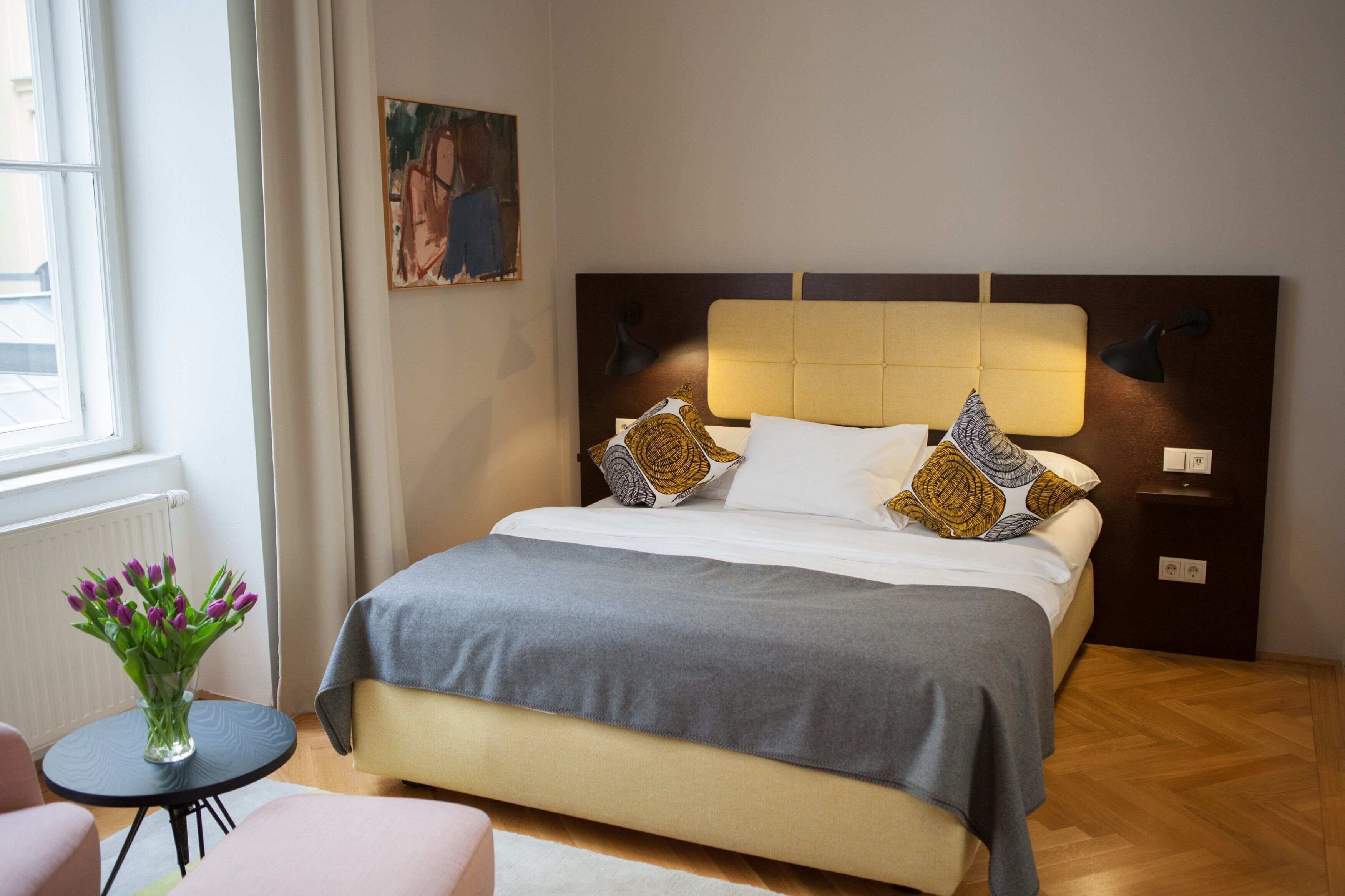 Ein ordentlich eingerichtetes Hotelzimmer mit Bett, Nachttischlampen, einem kleinen Tisch mit Blumenvase und einem Bild an der Wand.