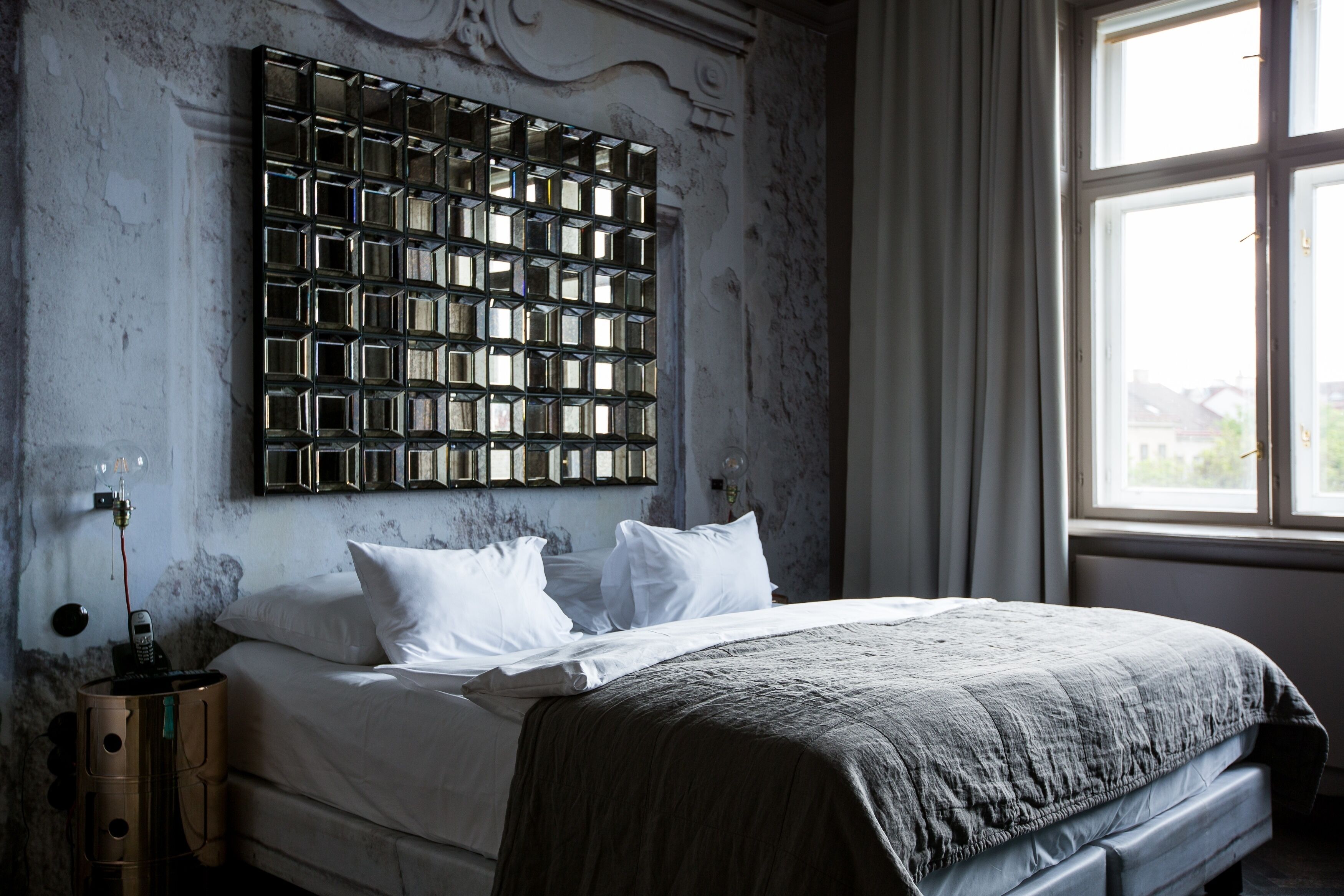 Een slaapkamer met een groot bed, een hoofdeinde met een geometrisch patroon, een nachtkastje met een lamp en een raam met gordijnen.