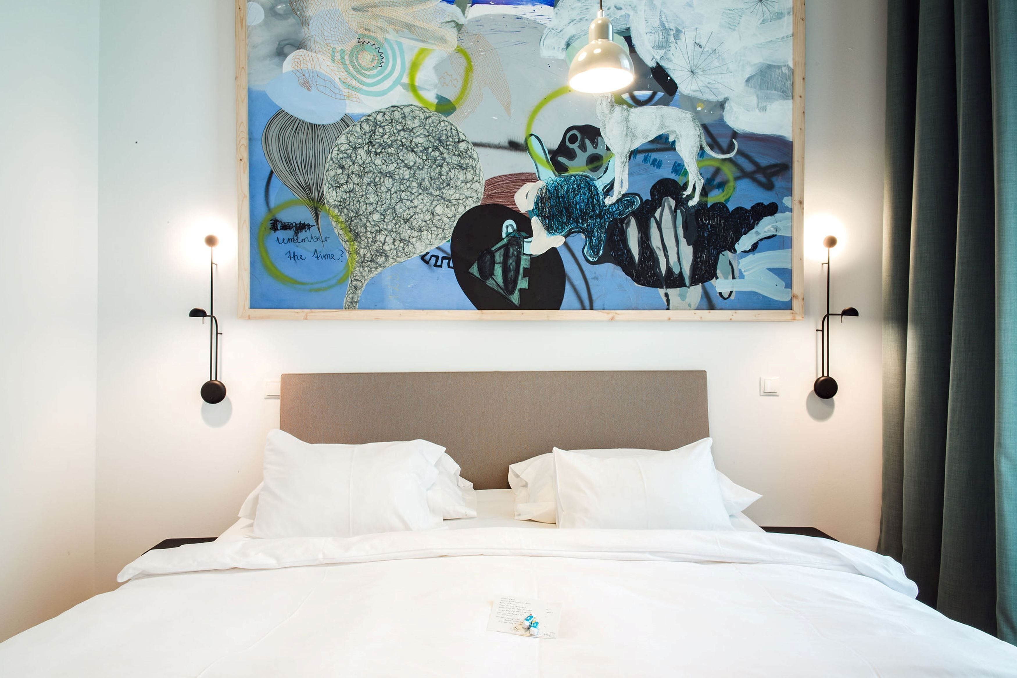 Quarto de hotel com cama, roupa de cama branca, um grande quadro abstrato na parede e duas luminárias de parede.