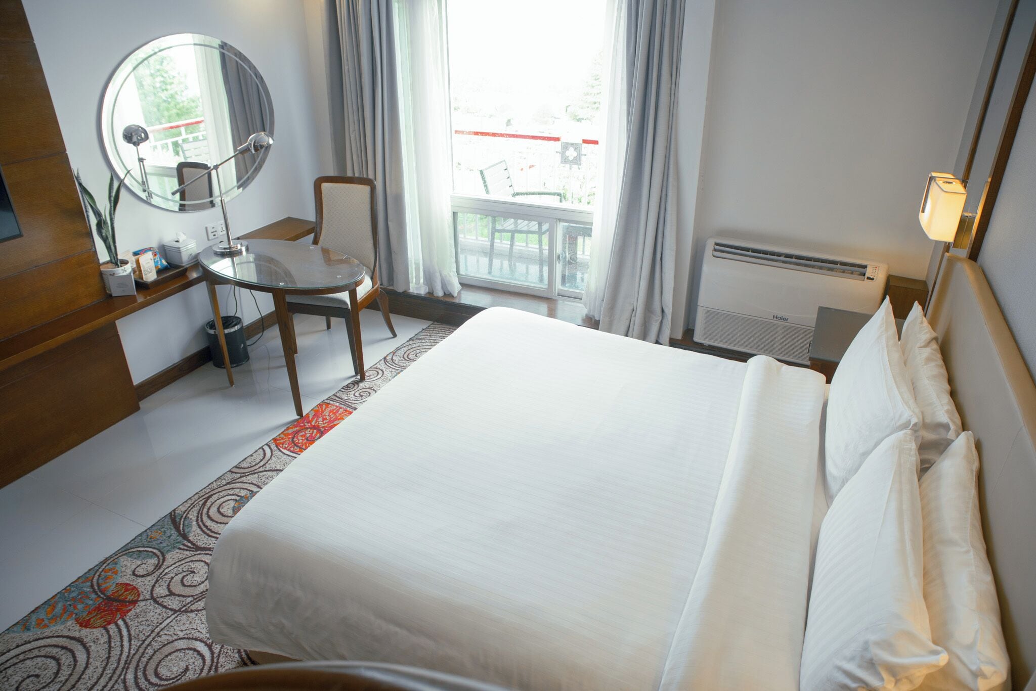 Een hotelkamer met een bed, een bureau met spiegel, een stoel, een raam met gordijnen en een radiator.