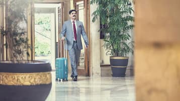 A man in a suit walking with a suitcase in a hotel lobby with marble floors and large potted plants.